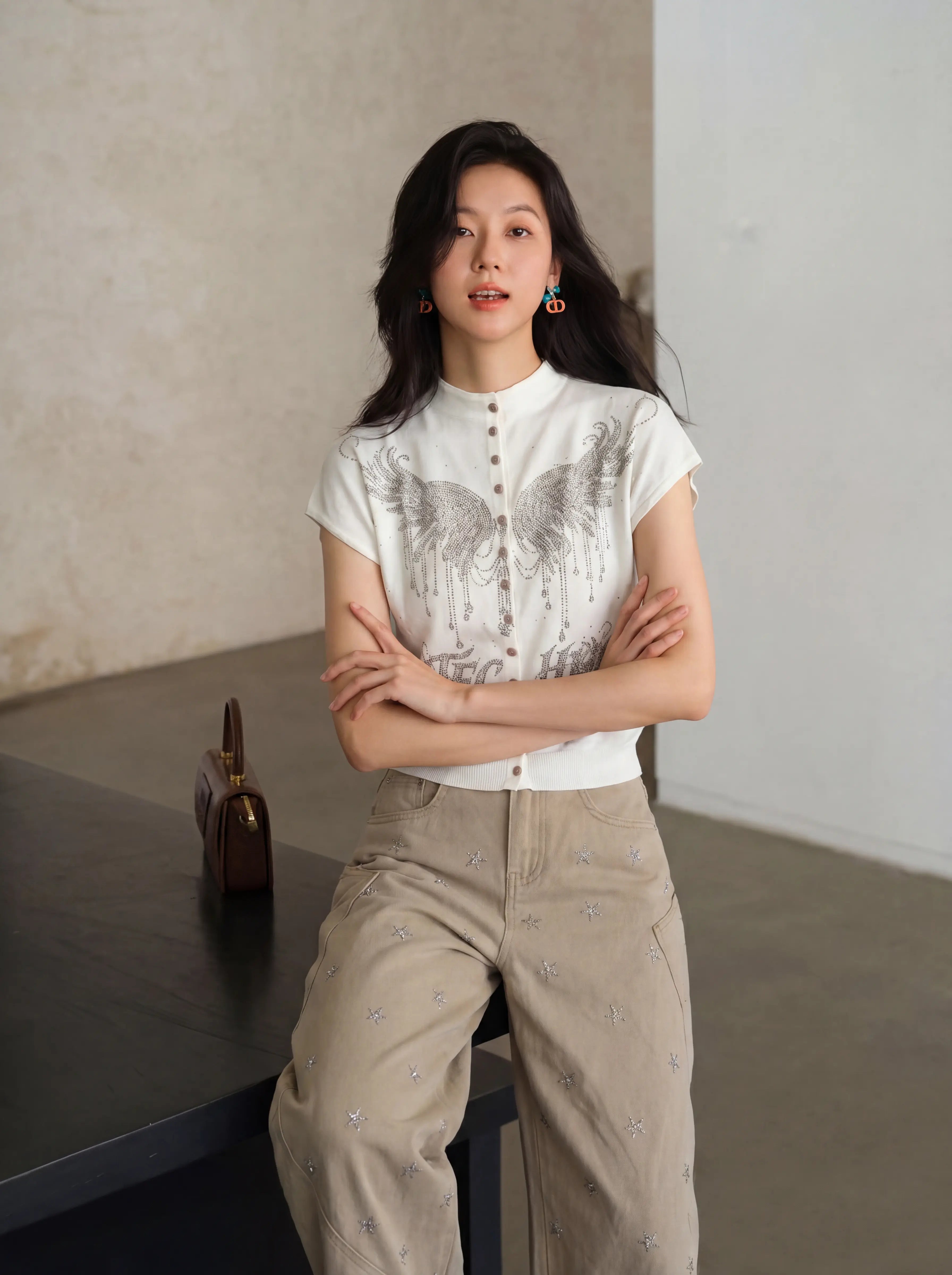 A woman stands indoors wearing a white short-sleeve button-front top with a gray wing graphic and beige wide-leg pants with small embroidered details. She is posed leaning against a dark table in a minimalist room with textured walls. TCH Rhinestone Phoenix Wings Button Knit Top