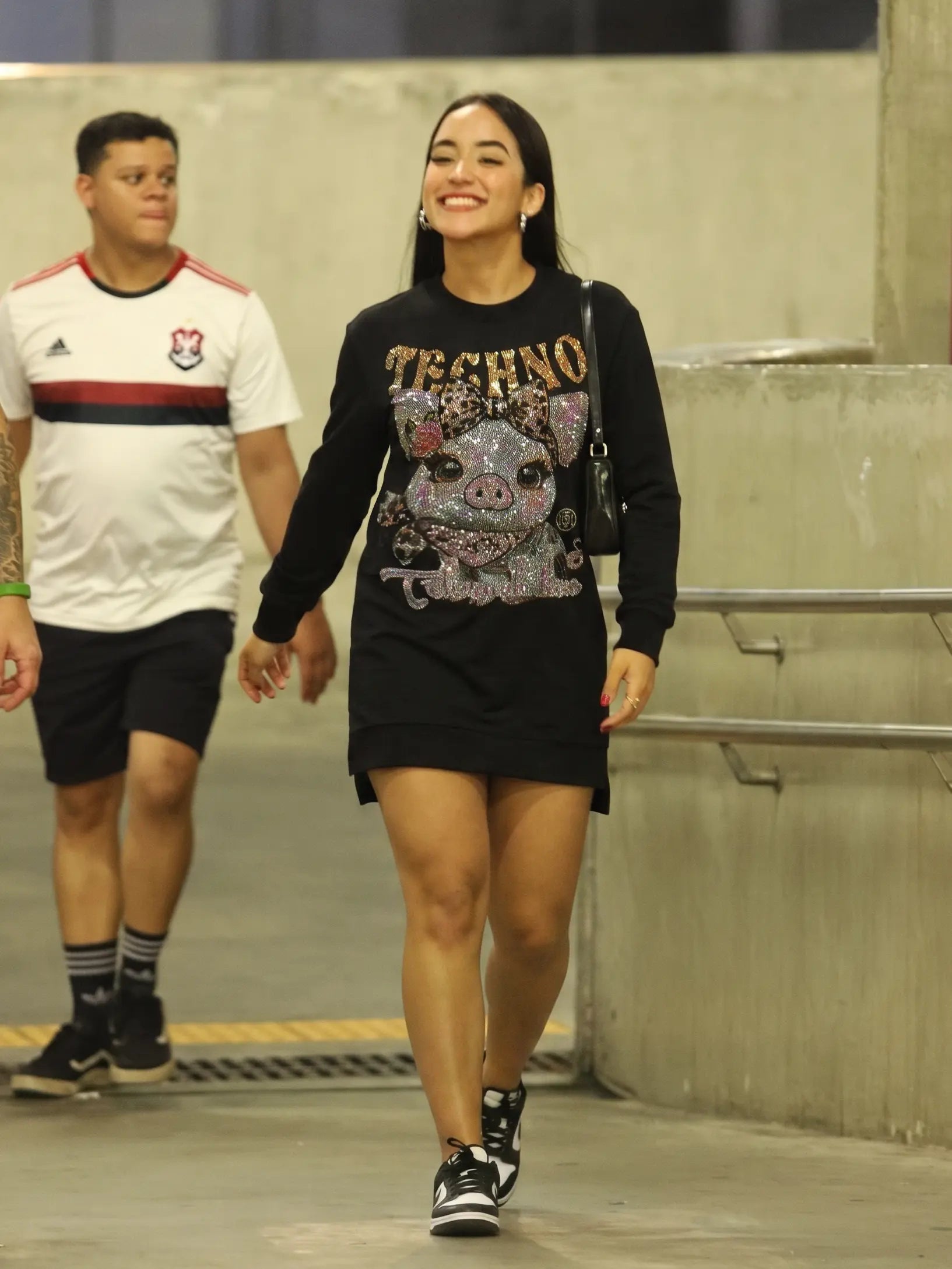 A woman walking in an indoor corridor wearing a black long-sleeve Techno sweatshirt dress with a large pink and silver rhinestone pig graphic on the front. A man in a white and black Flamengo jersey walks in the background. TCH Rhinestone Pig Graphic Long Sweatshirt Dress
