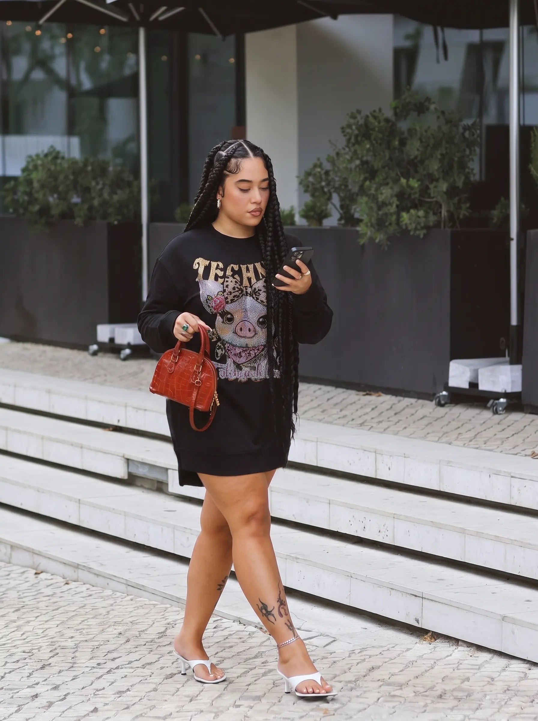 A woman walking down outdoor stone steps wearing a black graphic sweatshirt with a skull print and the word 'TECH' in gold, paired with a black mini skirt, white heeled sandals, and carrying a small red handbag. TCH Rhinestone Pig Graphic Long Sweatshirt Dress