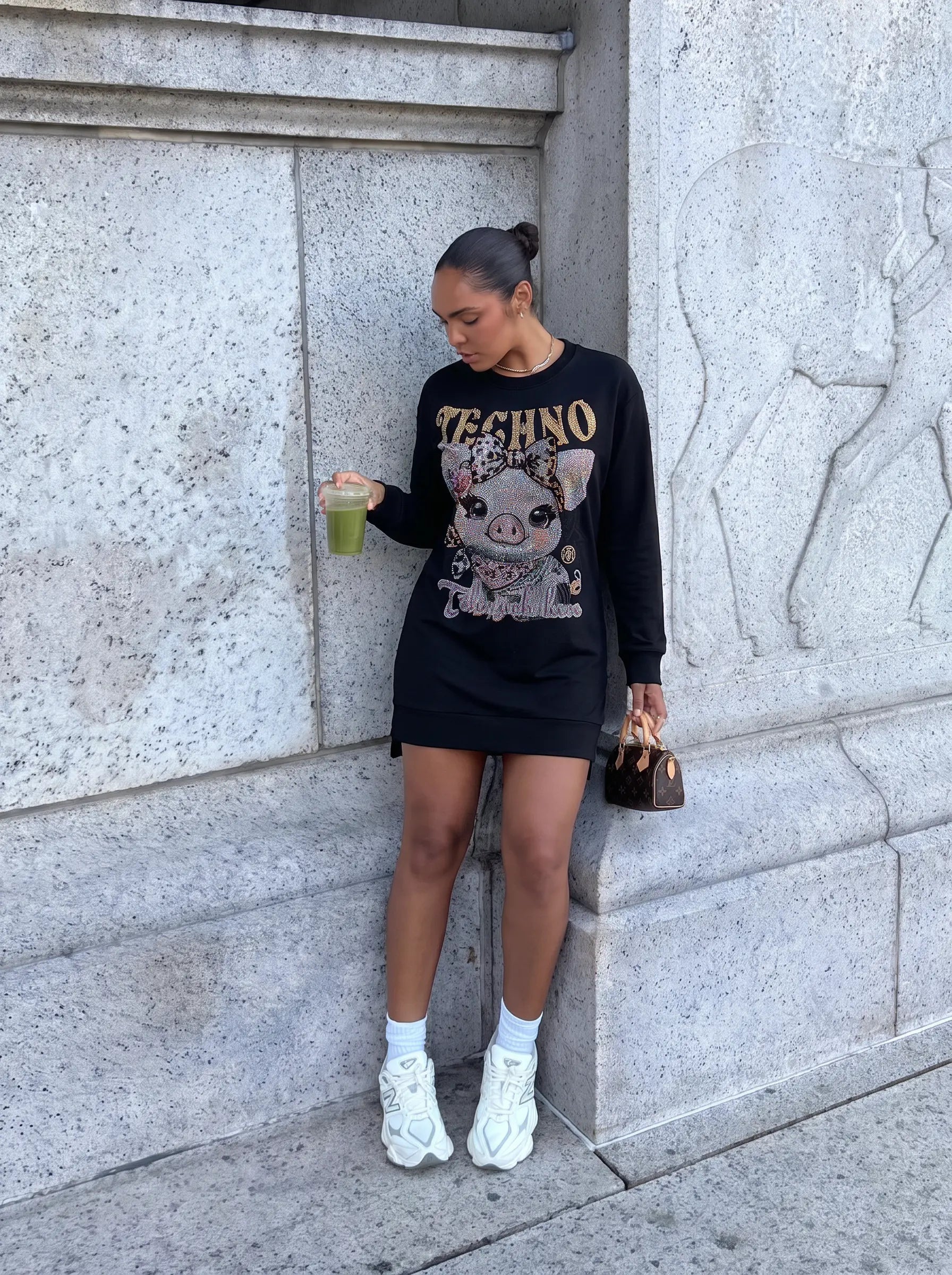 A woman stands against a gray stone wall wearing a black long-sleeve Techno sweatshirt dress with a pig graphic and gold lettering, paired with white sneakers and holding a green drink. TCH Rhinestone Pig Graphic Long Sweatshirt Dress