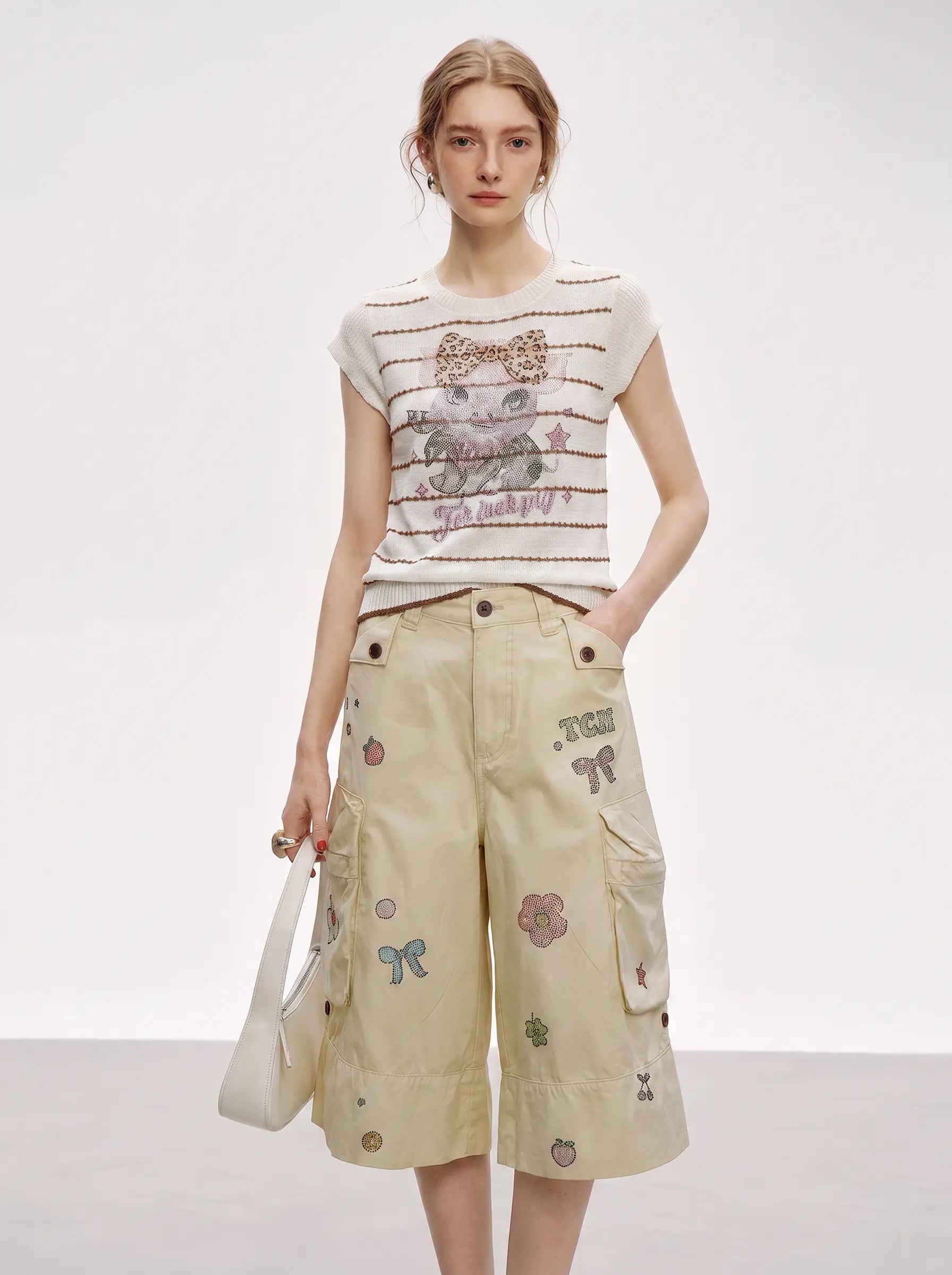 A model stands against a plain light gray studio background wearing a cream short-sleeve knit top with thin brown stripes and a central graphic print, paired with beige cargo shorts featuring colorful embroidered patches. She holds a white handbag. TCH Rhinestone Piggy Bow Striped Knit Top