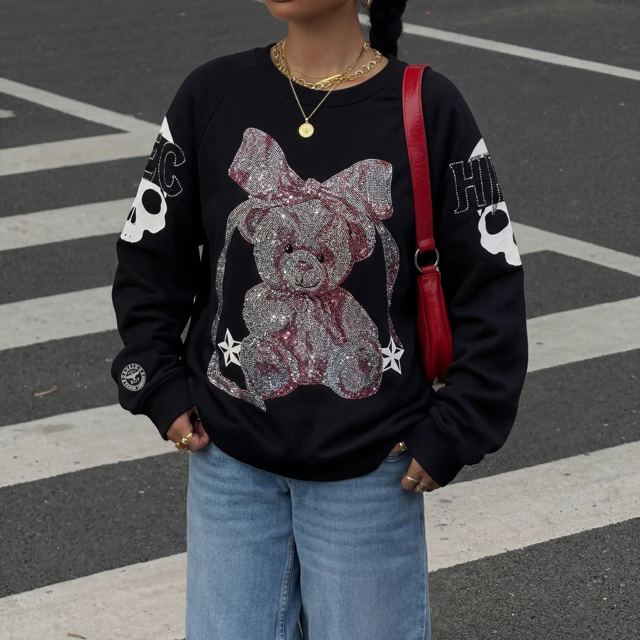 Person wearing a black sweatshirt with a teddy bear design, standing on a crosswalk. TCH Rhinestone Pink Bear Skull Sleeve Sweatshirt