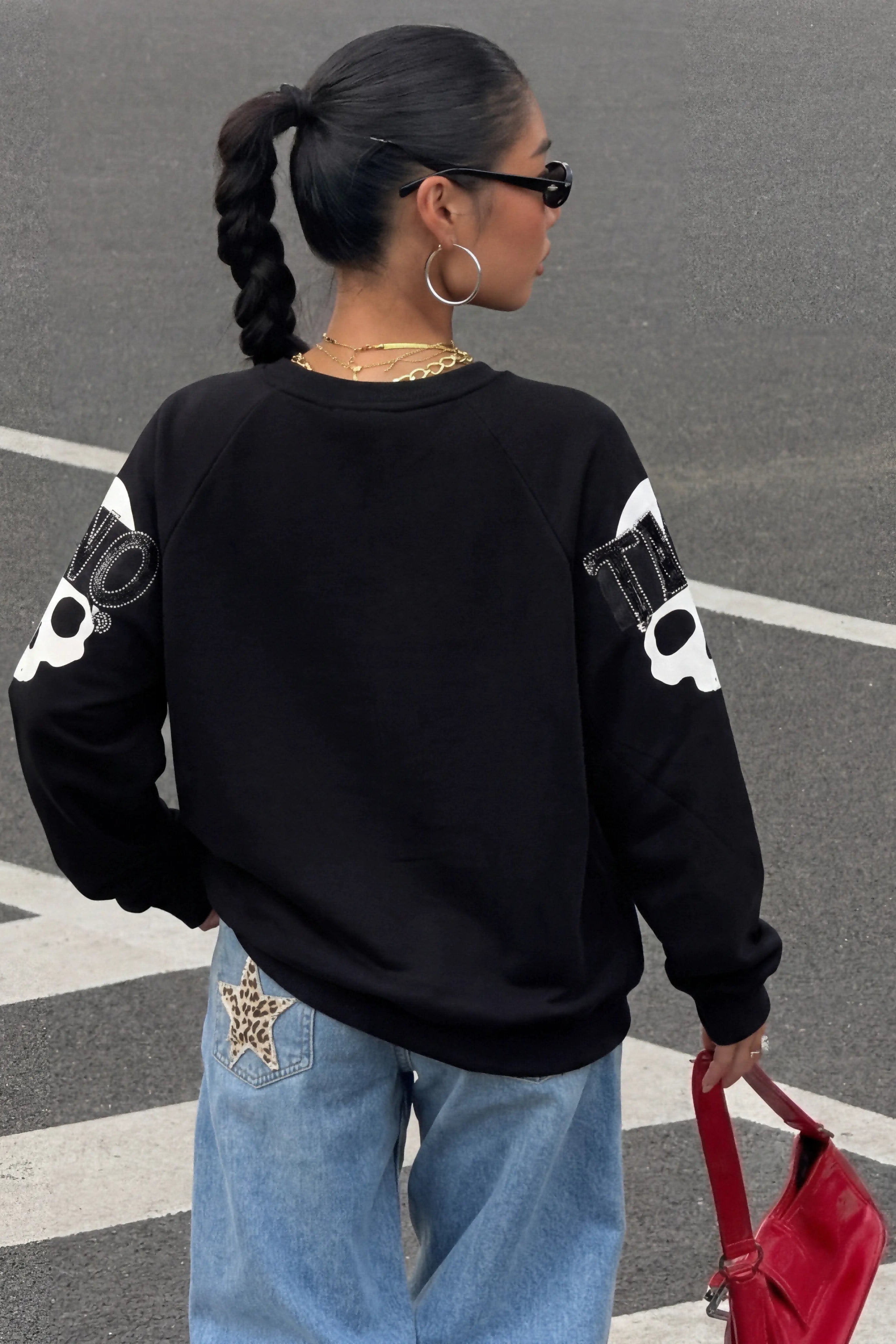 Person wearing a black sweatshirt with white designs, blue jeans, and a red bag on a street. TCH Rhinestone Pink Bear Skull Sleeve Sweatshirt