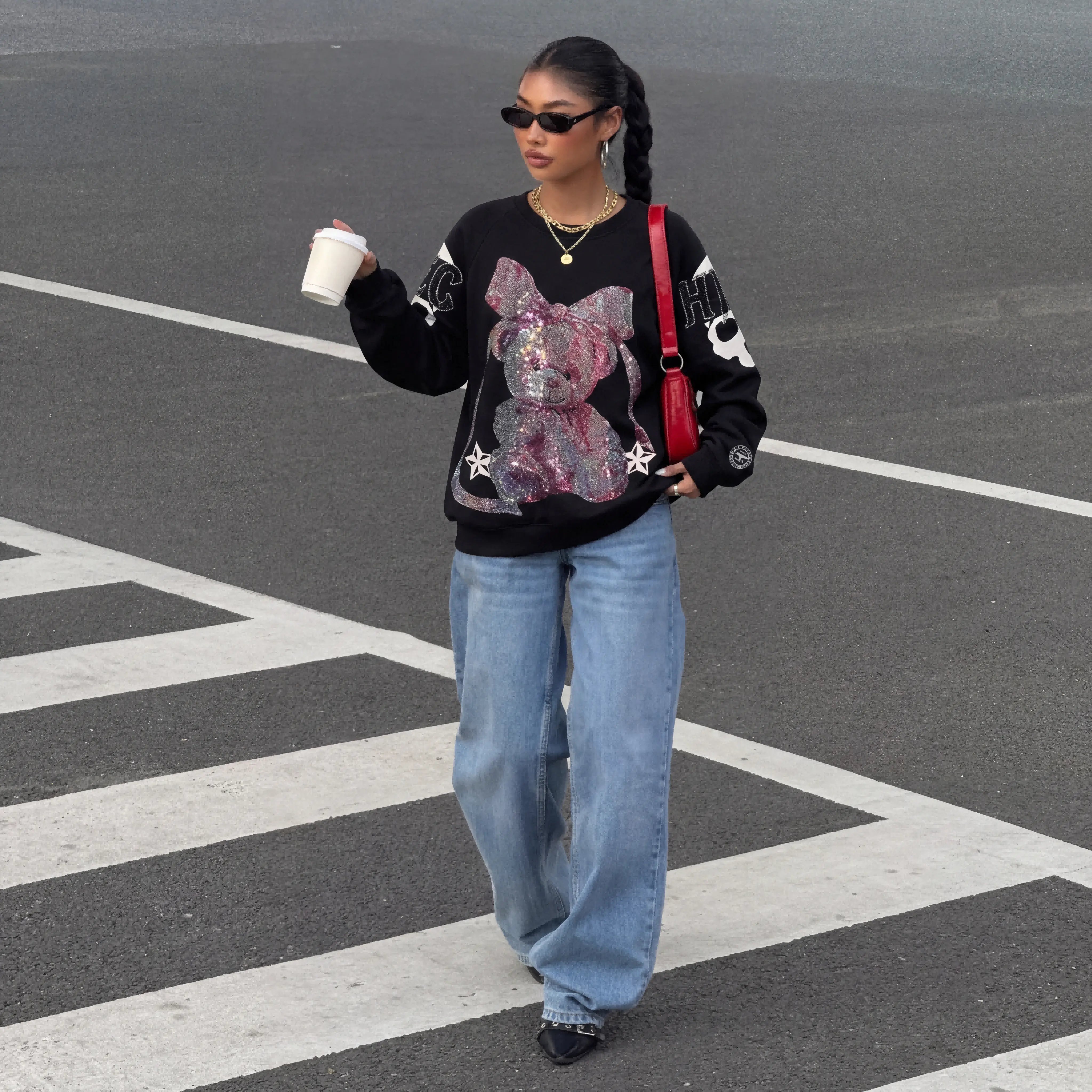 Woman in a black sweater with a pink bear design, blue jeans, and sunglasses walking on a street. TCH Rhinestone Pink Bear Skull Sleeve Sweatshirt