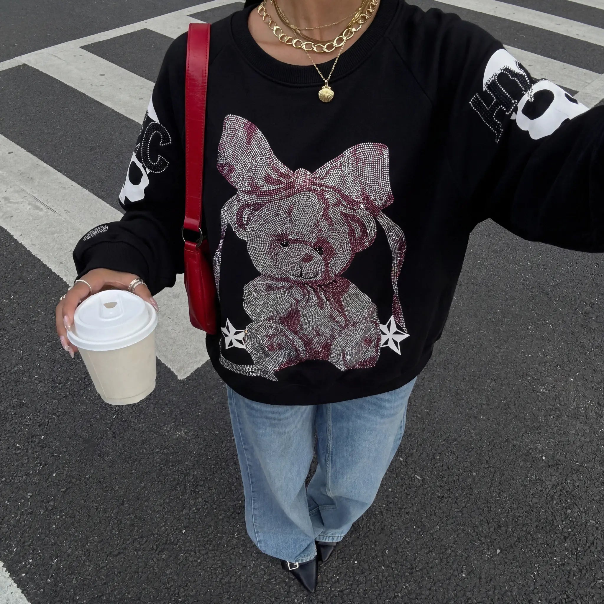 Person wearing a black sweater with a teddy bear design, holding a coffee cup on a street. TCH Rhinestone Pink Bear Skull Sleeve Sweatshirt