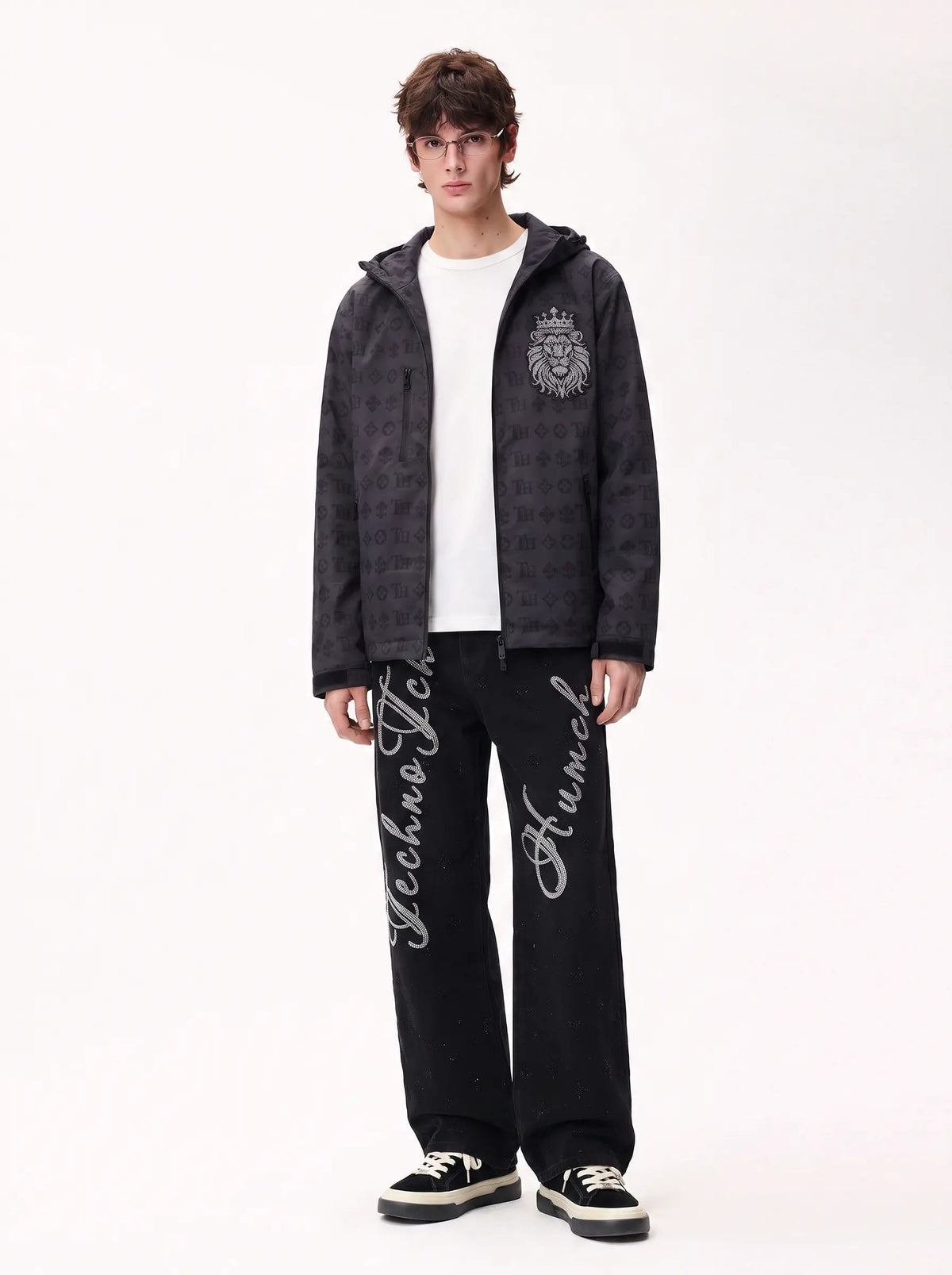 A male model stands against a plain white studio background wearing a dark patterned zip-up hoodie with a silver crest on the chest, a white t-shirt, black sweatpants with white script embroidery, and black sneakers. TCH Rhinestone Script Graphic Wide-Leg Jeans