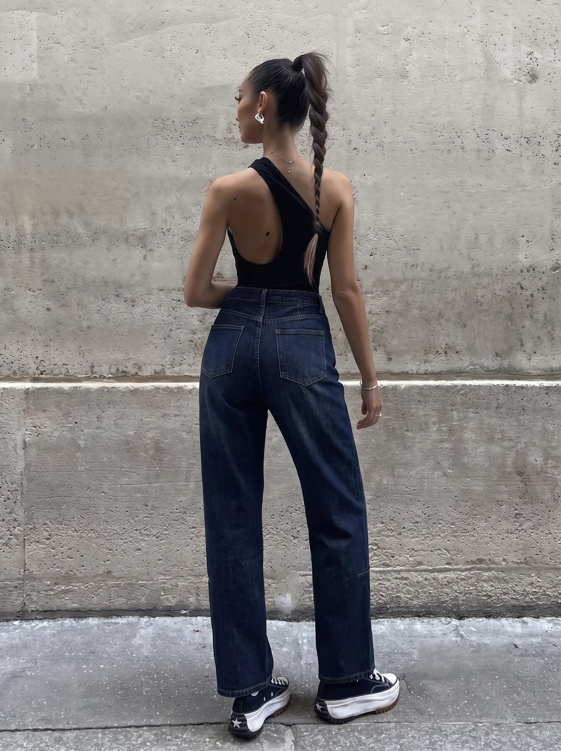 A woman stands with her back to the camera wearing a black one-shoulder backless top, dark blue wide-leg jeans, and black platform sneakers, posing against a textured concrete wall. TCH Rhinestone Shooting Star Galaxy Jeans