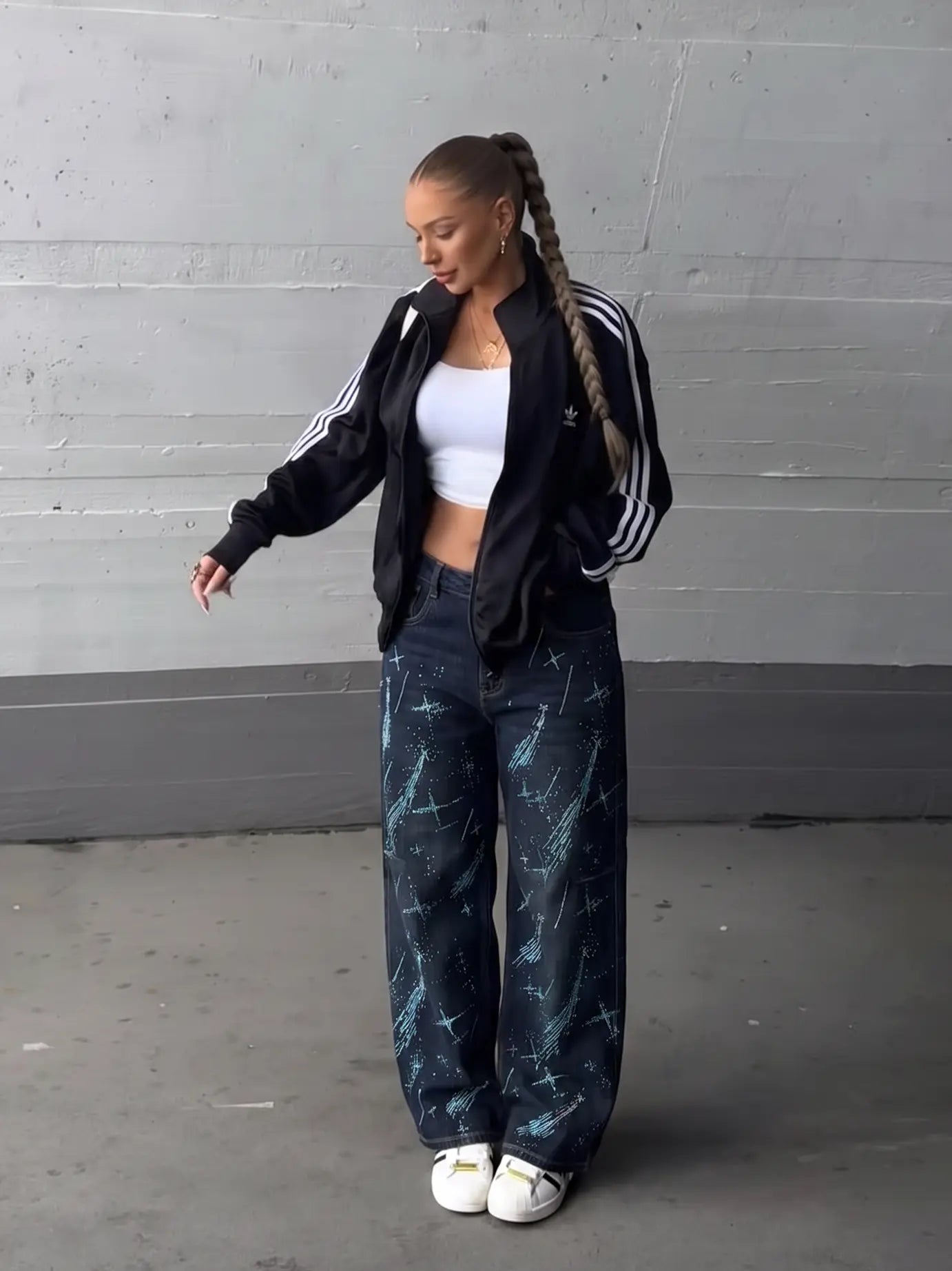 A woman stands in a studio wearing a black Adidas track jacket with white stripes, a white crop top, dark blue wide-leg jeans with a blue marble pattern, and white sneakers. TCH Rhinestone Shooting Star Galaxy Jeans