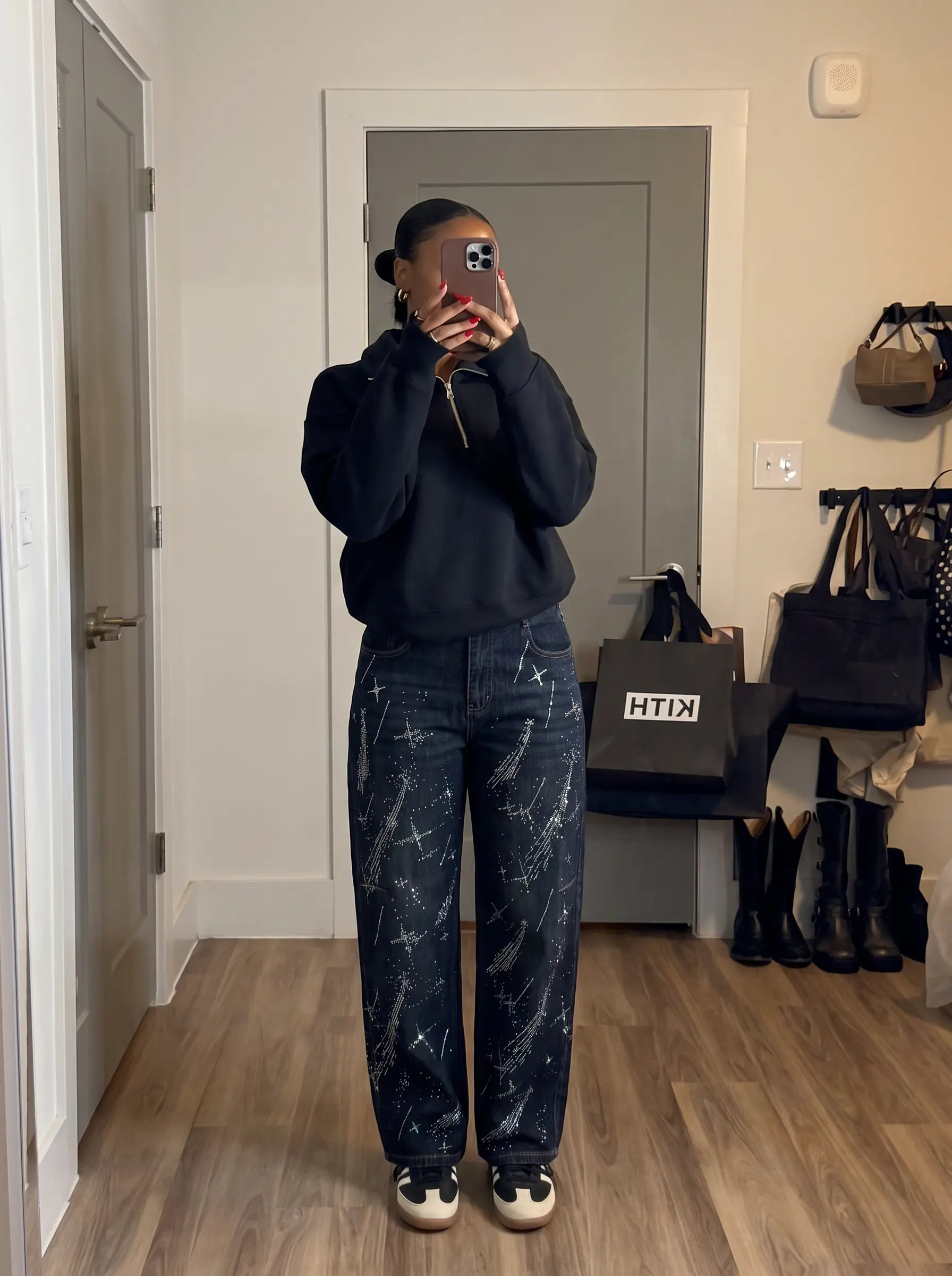A person taking a mirror selfie in a home hallway, wearing a black crewneck sweatshirt, dark blue jeans with a white abstract pattern, and black and white sneakers. A black bag with the white text 'HTIN' is visible on a shelf in the background. TCH Rhinestone Shooting Star Galaxy Jeans