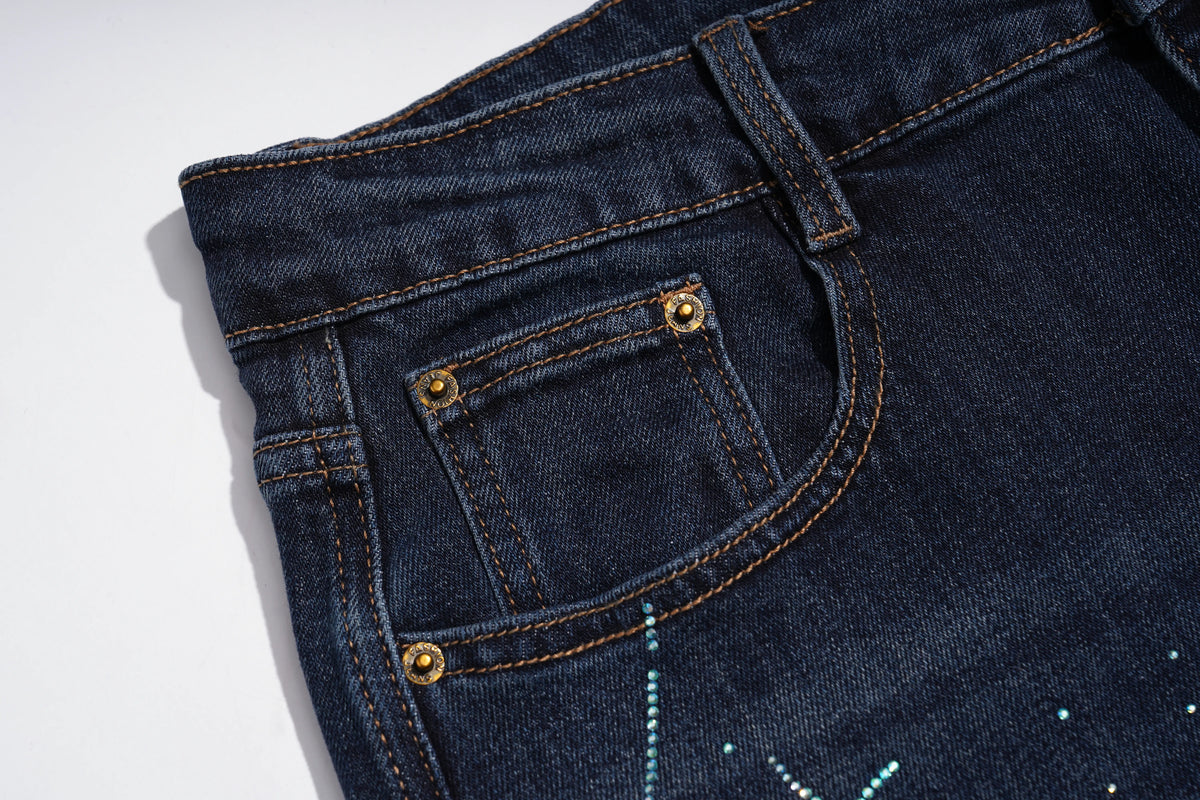 Close-up of a dark indigo denim pair of jeans showing the back pocket with gold-tone rivets and contrast stitching on a white background. TCH Rhinestone Shooting Star Galaxy Jeans