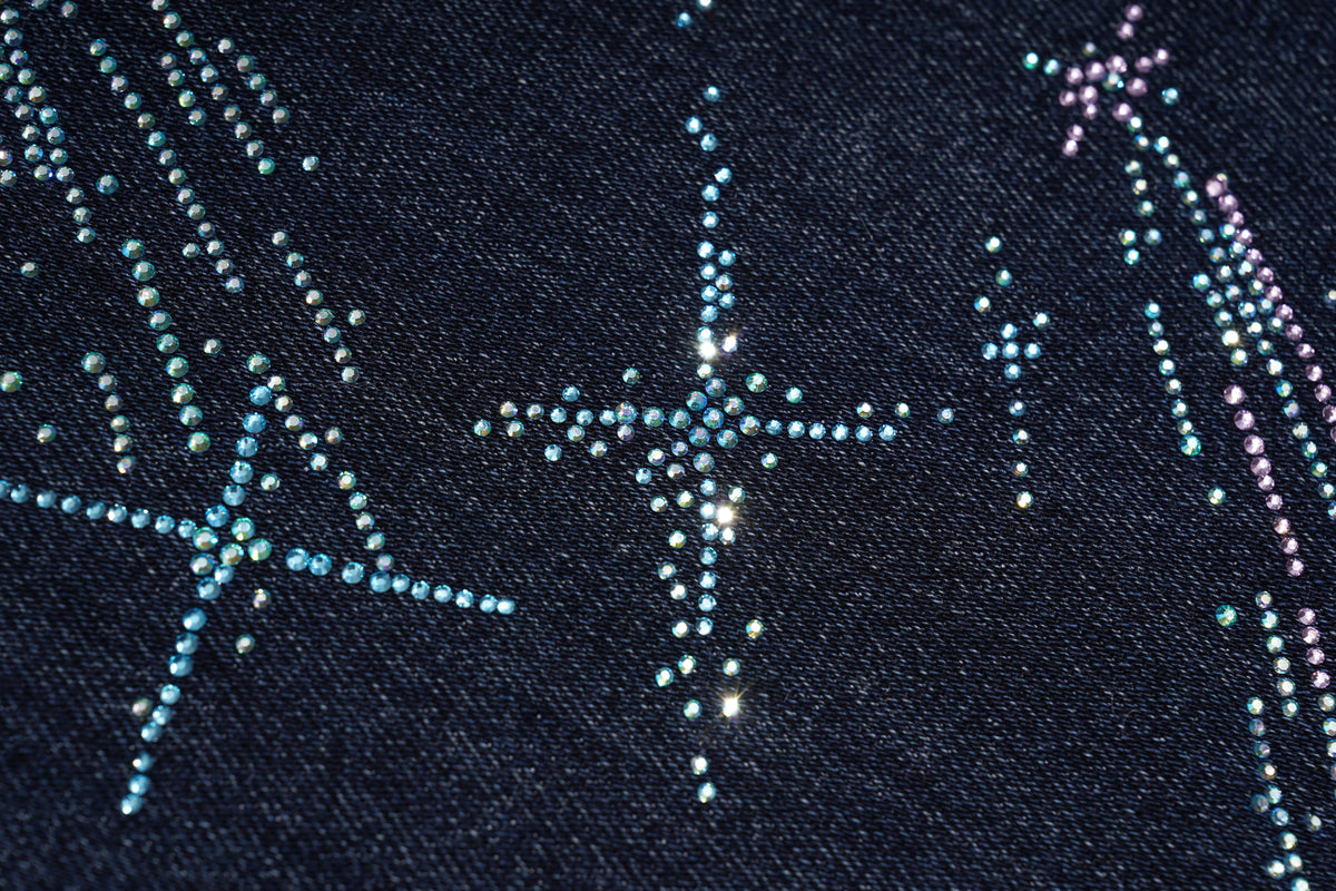 Close-up of a dark navy fabric with decorative rhinestone star and shooting star patterns in light blue, silver, and pink. TCH Rhinestone Shooting Star Galaxy Jeans