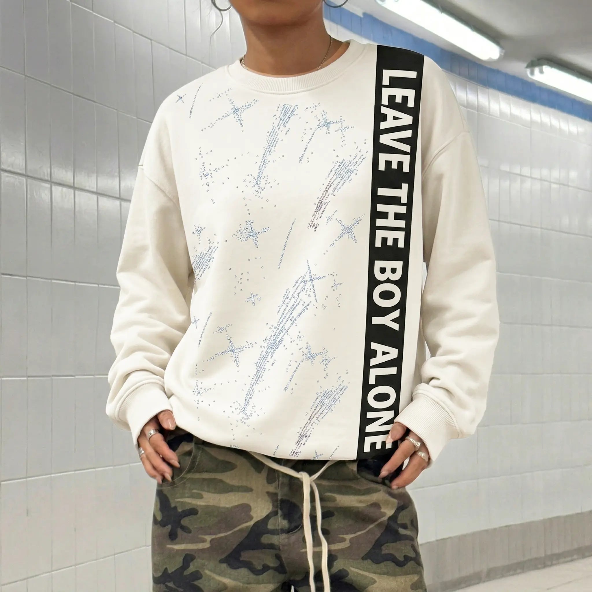 Person wearing a white sweatshirt with text and camouflage pants in a tiled room. TCH Rhinestone Shooting Star Sweatshirt