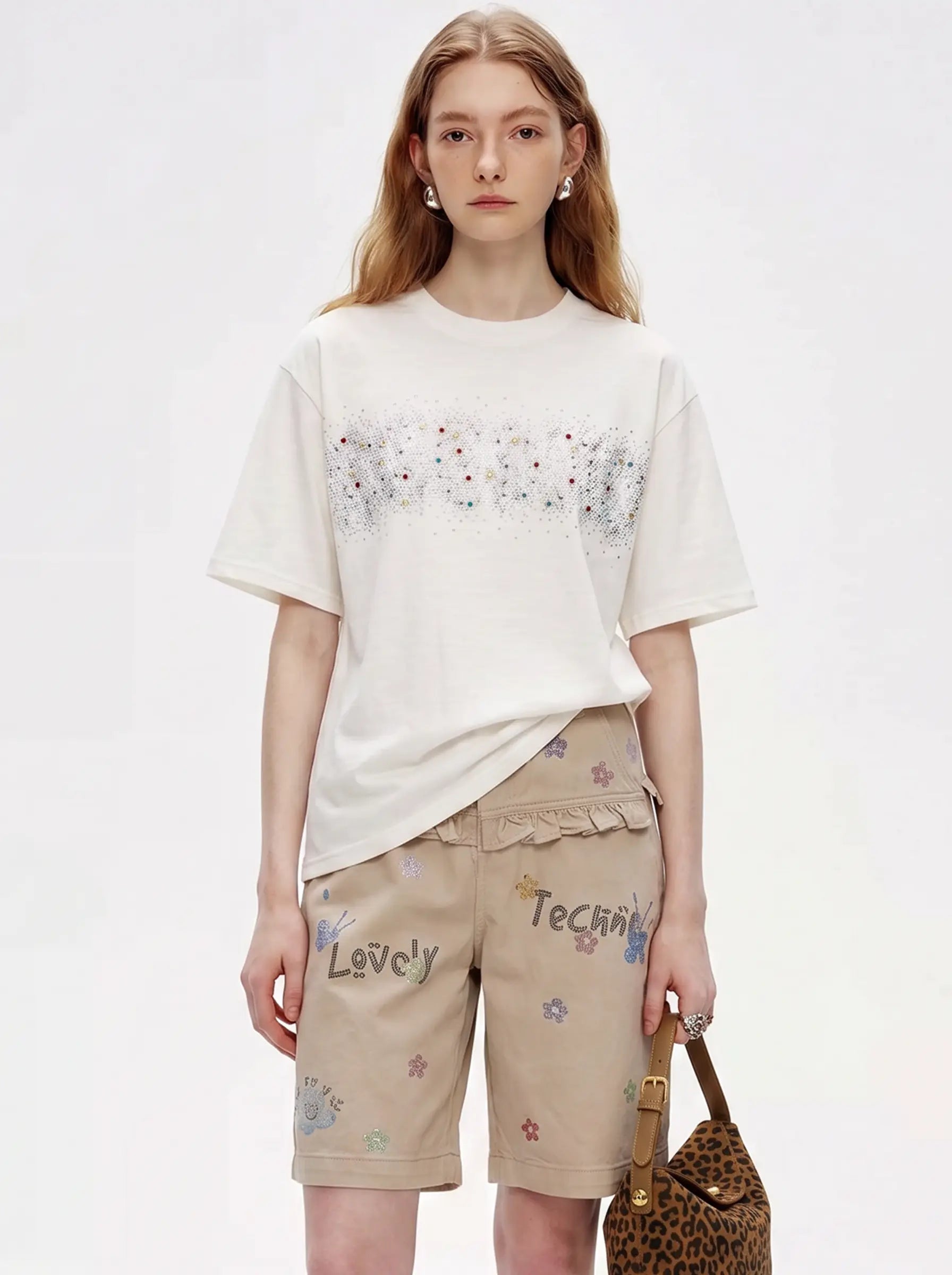 A model wearing a cream-colored short-sleeve t-shirt with a horizontal band of colorful embroidered flowers, paired with beige shorts featuring embroidered text and floral details, holding a leopard-print handbag against a plain white background. TCH Rhinestone Sparkle Crystal Graphic White T-Shirt