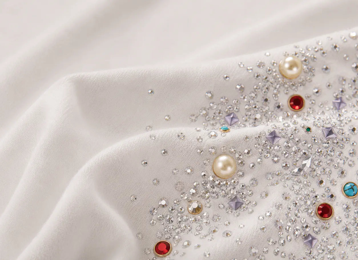 Close-up of a white or ivory fabric surface embellished with scattered clear rhinestones, small pearl-like beads, and a few colored gem accents including red, blue, and lavender. TCH Rhinestone Sparkle Crystal Graphic White T-Shirt