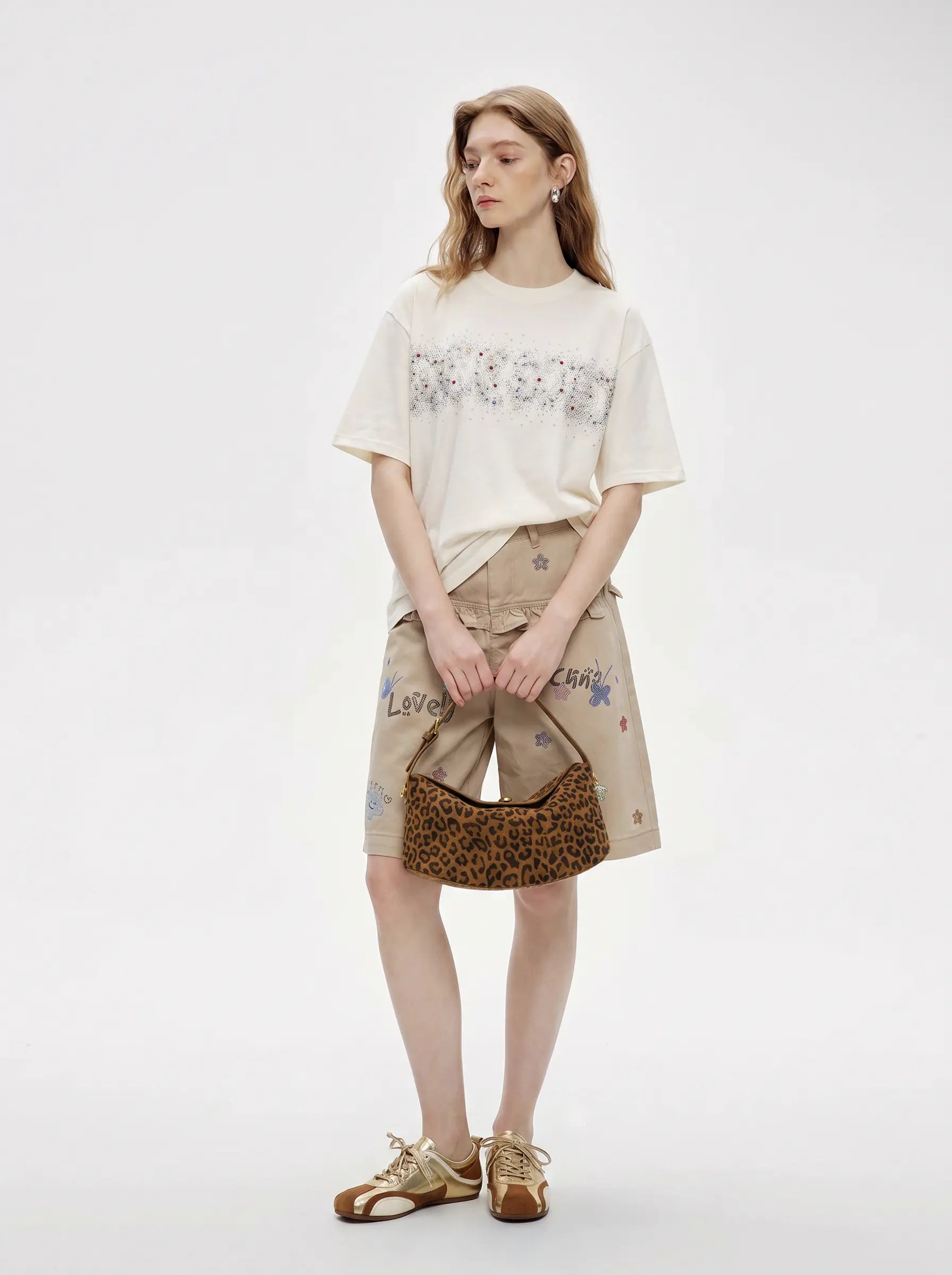 A model stands against a light gray studio background wearing a cream graphic T-shirt, beige shorts with colorful text and star prints, a leopard-print shoulder bag, and metallic gold sandals. TCH Rhinestone Sparkle Crystal Graphic White T-Shirt
