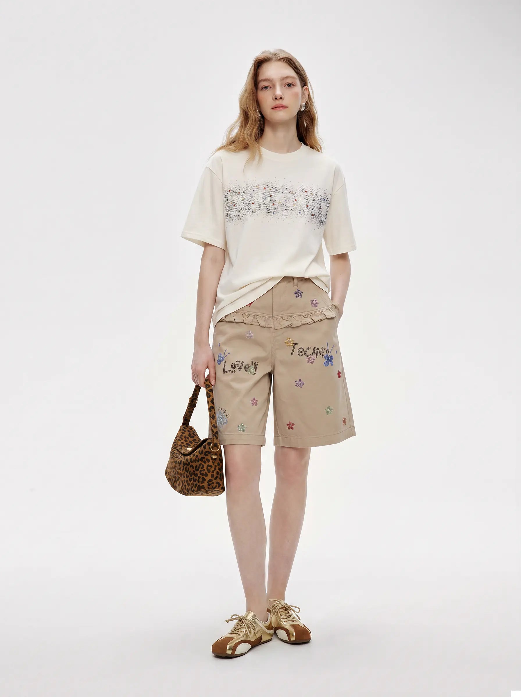 A model stands against a light gray studio background wearing a cream graphic t-shirt, beige embroidered shorts, and brown sandals, while holding a brown patterned handbag. TCH Rhinestone Sparkle Crystal Graphic White T-Shirt