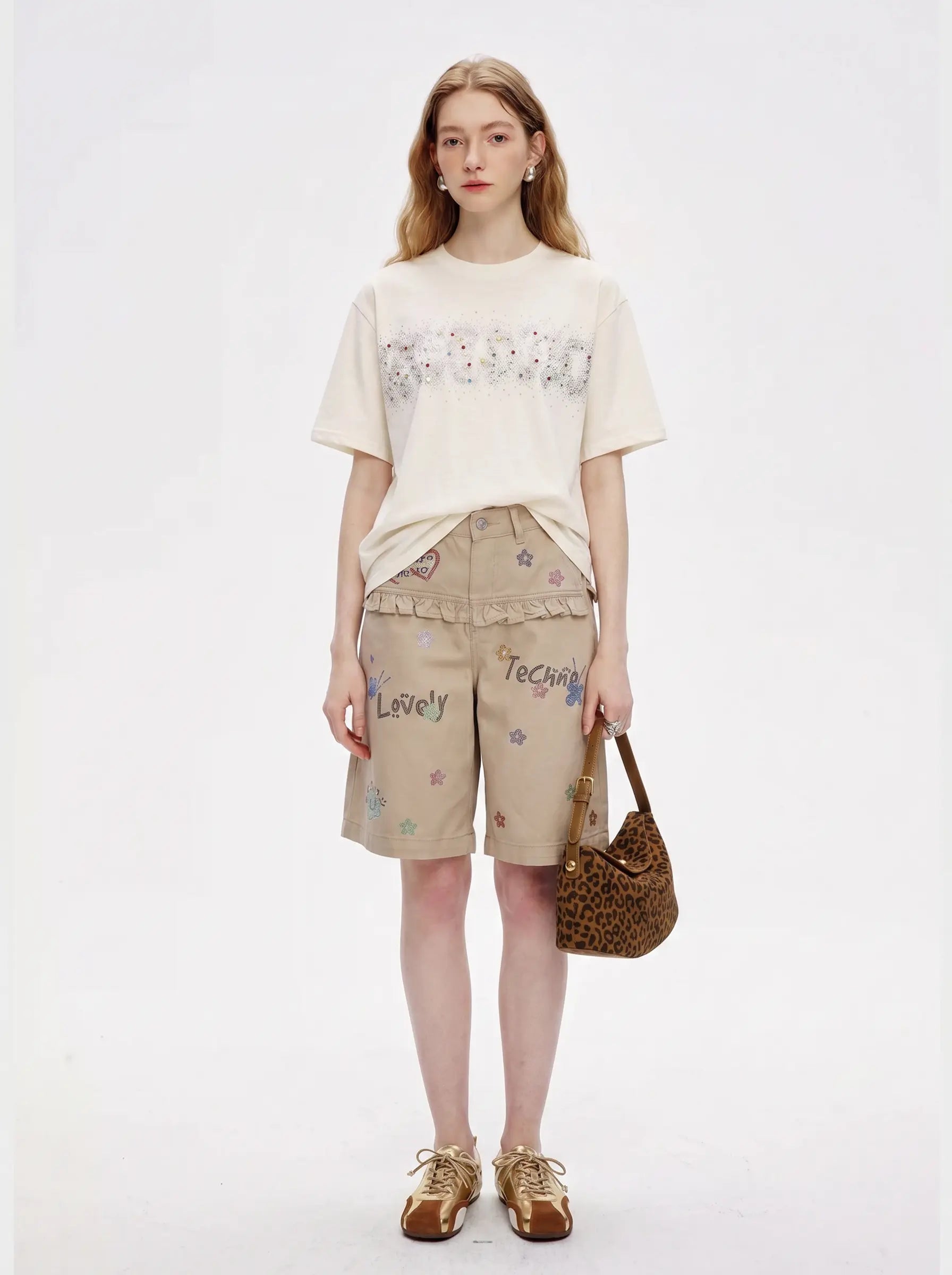 A model stands against a plain white studio background wearing a cream t-shirt with a floral pattern, beige shorts with embroidered details, and brown shoes, while holding a brown patterned handbag. TCH Rhinestone Sparkle Crystal Graphic White T-Shirt