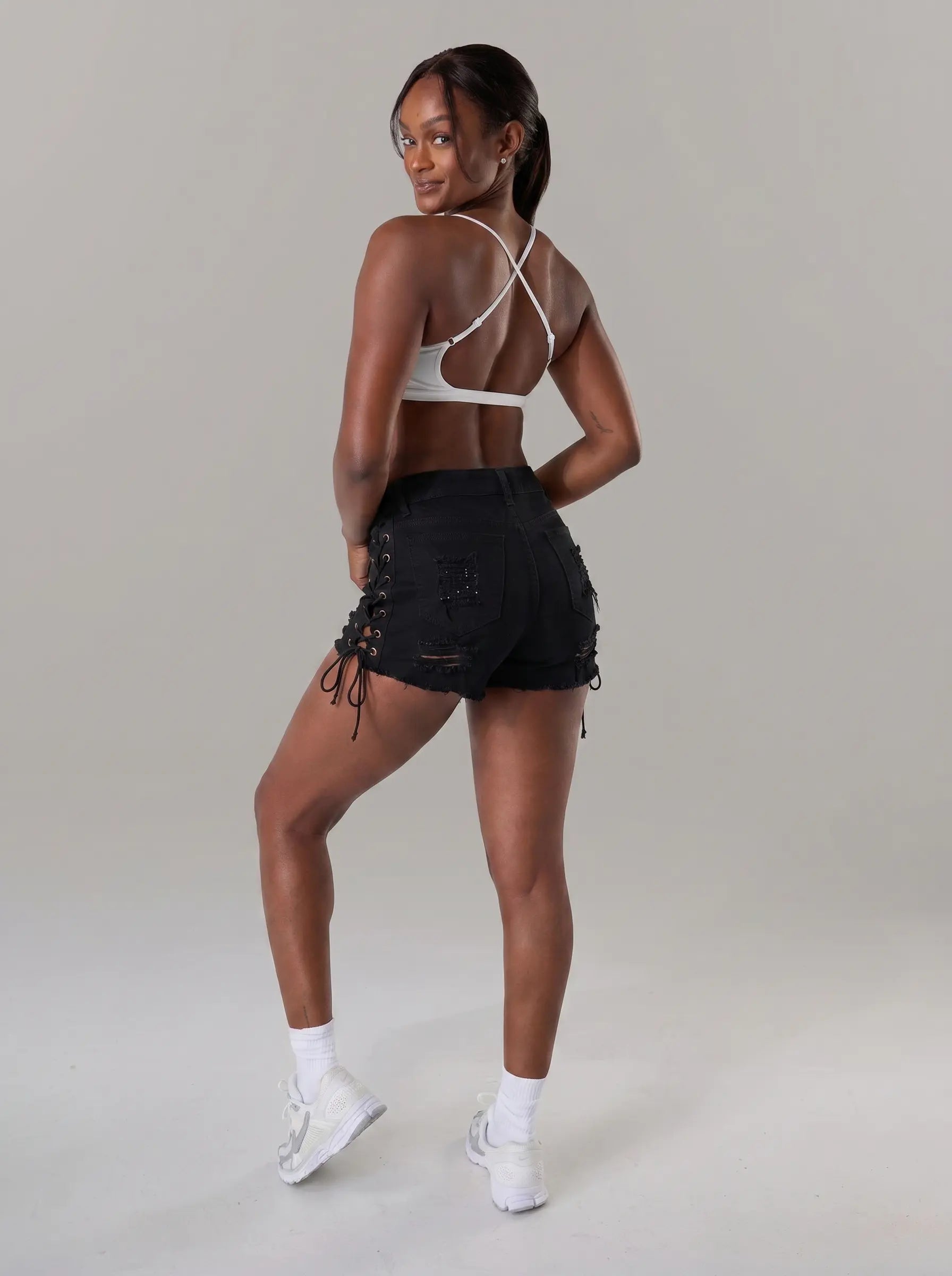 A woman posing from behind wearing black lace-up denim shorts, a white sports bra, and white sneakers against a neutral gray studio background. TCH Rhinestone Sunflower Distressed Denim Shorts