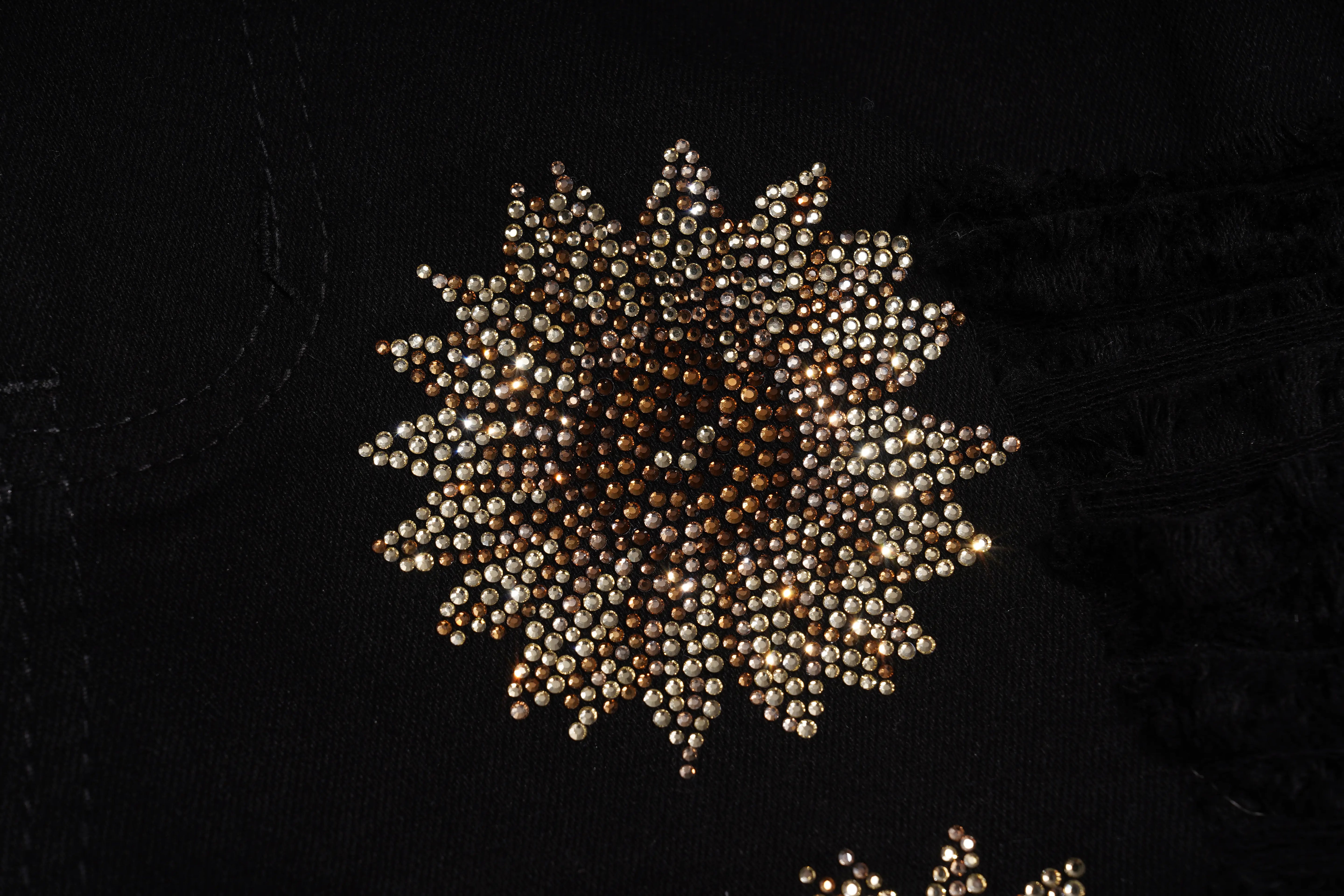 A close-up of a circular embellished brooch or decorative applique made of many small metallic beads, photographed on a black fabric background. TCH Rhinestone Sunflower Distressed Denim Shorts