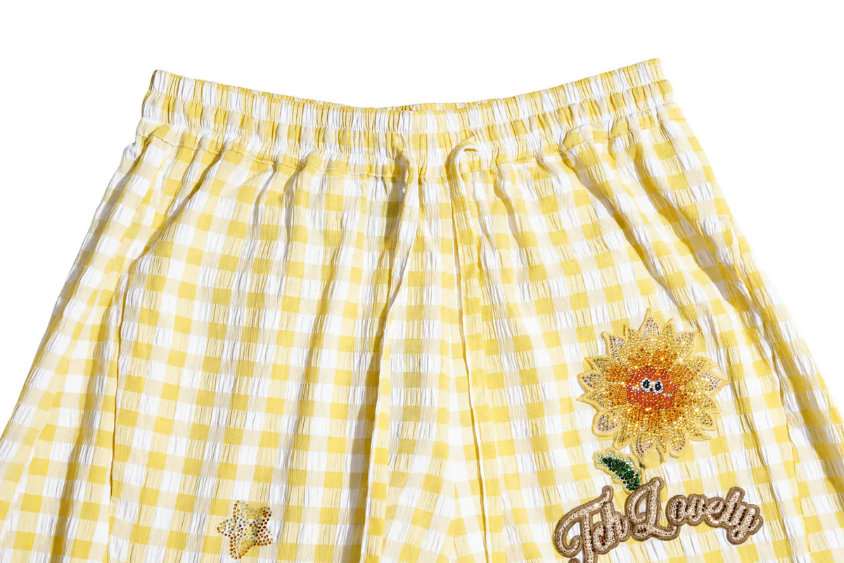 Yellow and white gingham checkered shorts featuring a large embroidered sunflower with 'Fits Lovely' text on the right leg and a small gold glitter star on the left leg, shown against a plain white background. TCH Rhinestone Sunflower Gingham Cargo Wide-Leg Pants