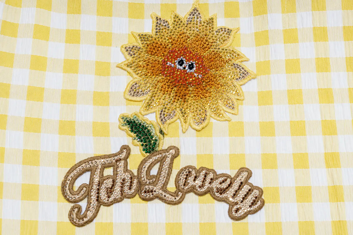 A close-up of a yellow and white gingham fabric featuring an embroidered sunflower patch with a smiling face and the phrase 'Fh Lovely' in cursive script below it. TCH Rhinestone Sunflower Gingham Cargo Wide-Leg Pants