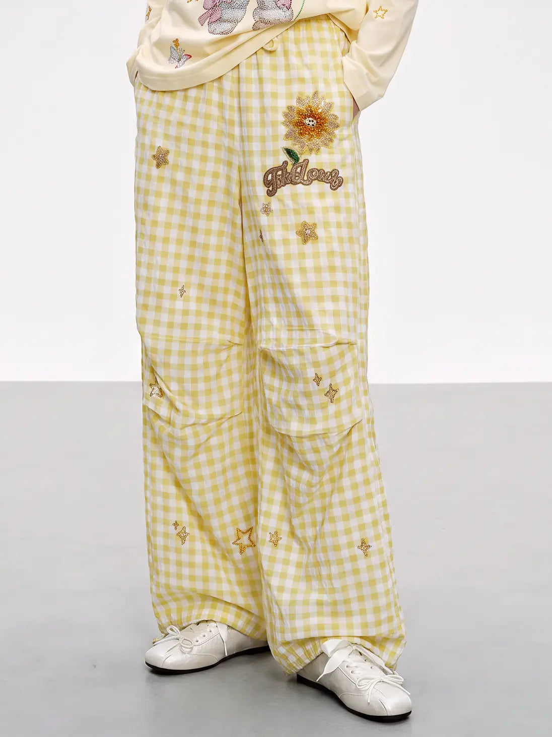 A person wearing wide-leg yellow and white gingham cargo pants with embroidered flowers, stars, and a 'Goldilocks' patch, paired with a cream sweatshirt and white sneakers. TCH Rhinestone Sunflower Gingham Cargo Wide-Leg Pants