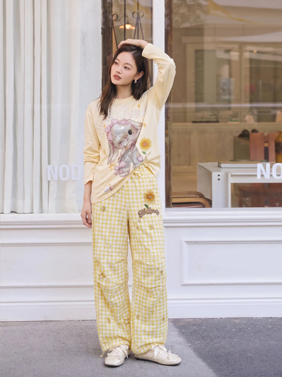 A woman stands in front of a storefront window wearing a light yellow long-sleeve top with a bunny and sunflower graphic, paired with matching yellow gingham pants and white sneakers. TCH Rhinestone Sunflower Gingham Cargo Wide-Leg Pants
