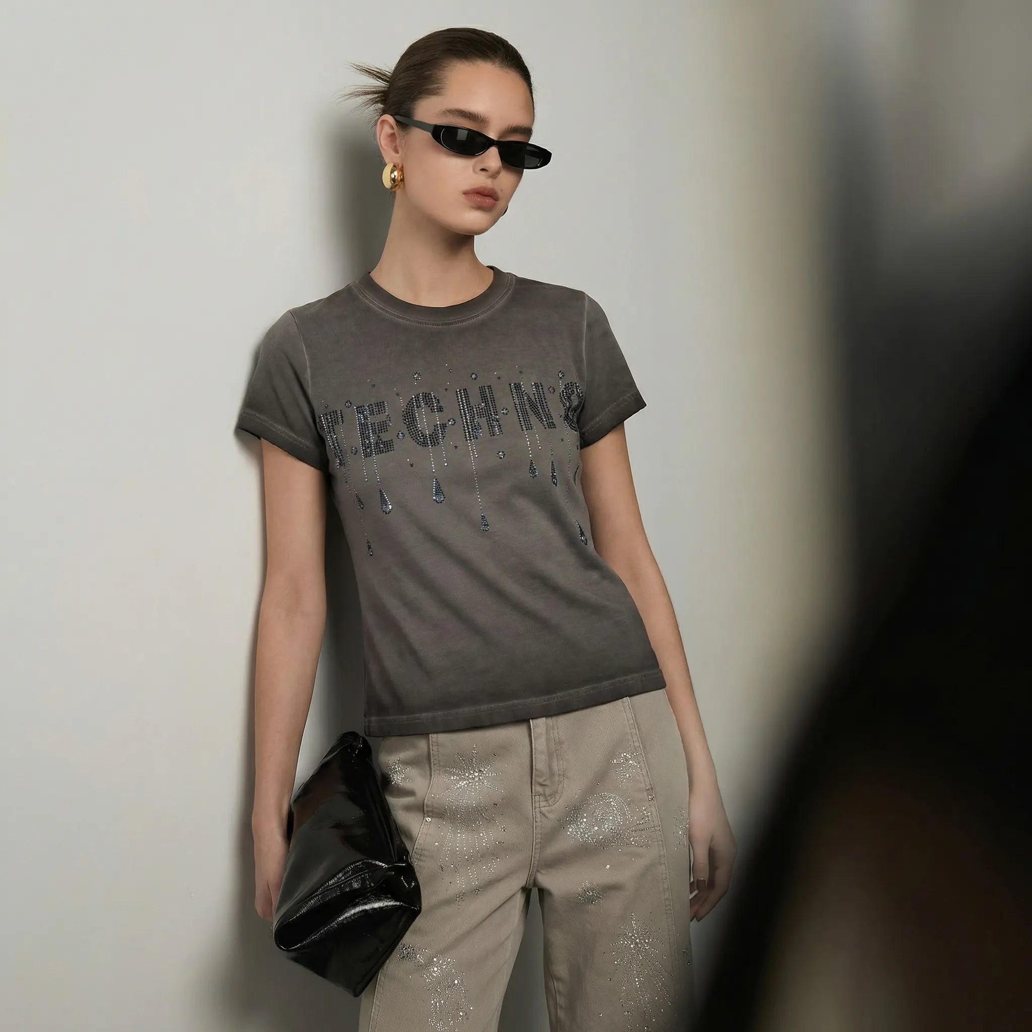 A model wearing a dark gray graphic t-shirt with 'TECHING' printed across the chest in a distressed, dripping font, paired with beige pants and holding a black clutch bag. TCH Rhinestone TECHNO Graphic Washed T-Shirt