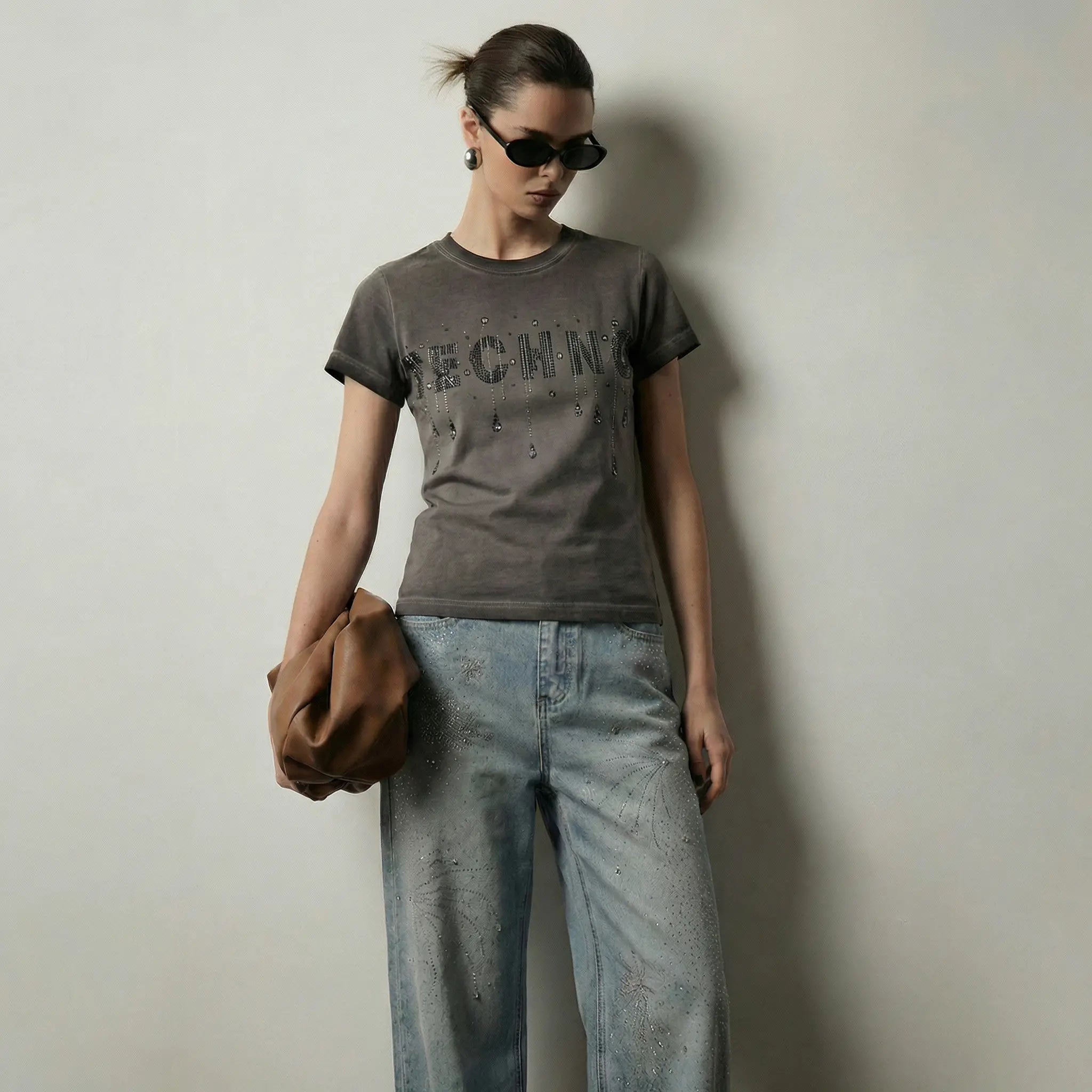 A model wearing a dark gray graphic t-shirt with 'SECHN' printed on the front, paired with light-wash distressed jeans and holding a brown leather hat against a plain light gray wall. TCH Rhinestone TECHNO Graphic Washed T-Shirt