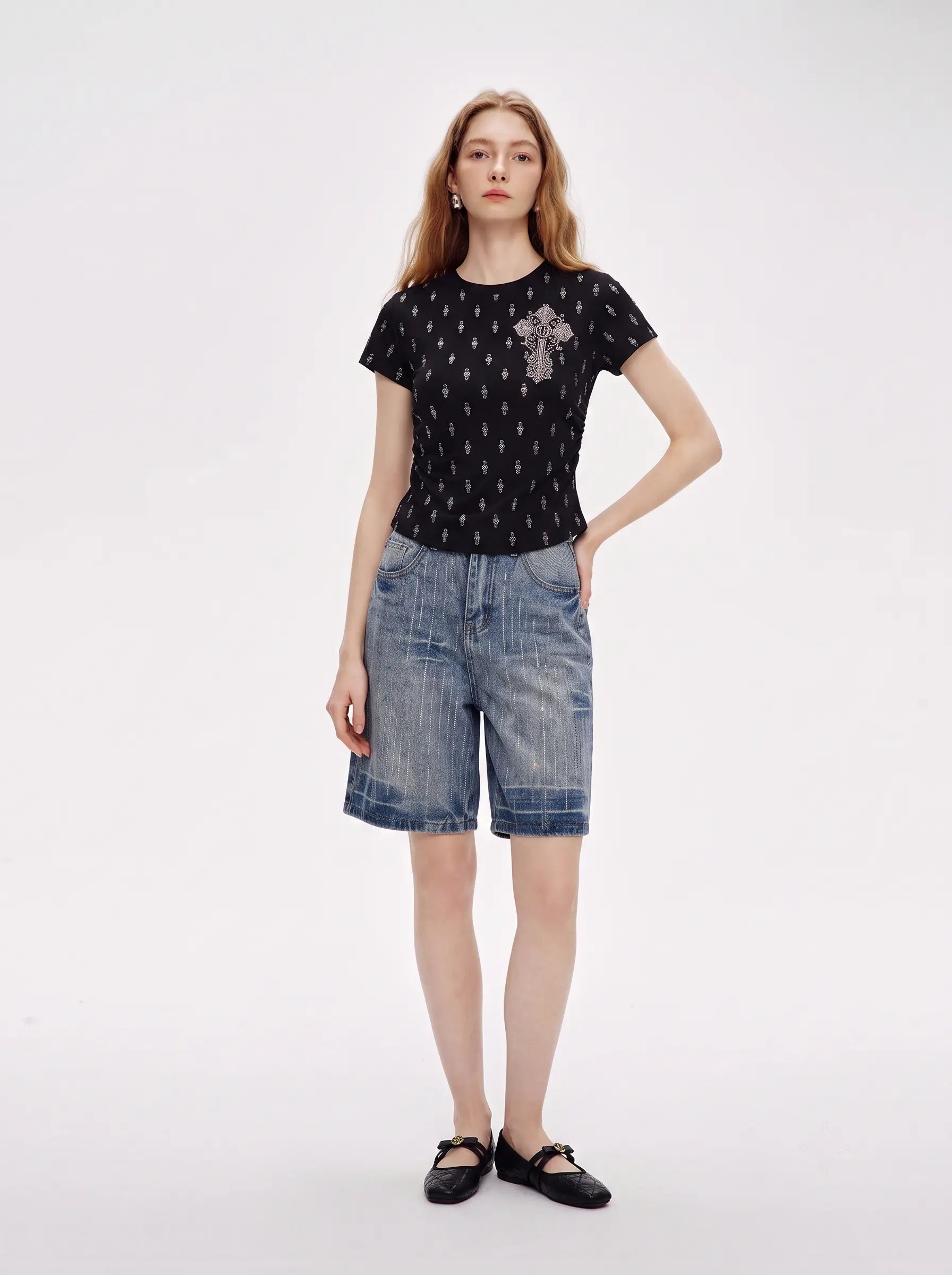 A model stands against a light gray studio background wearing a black short-sleeve top with a small repeating pattern and a silver-toned cross motif on the chest, paired with blue denim shorts and black flats. TCH Rhinestone Techno Cross Graphic Black T-Shirt