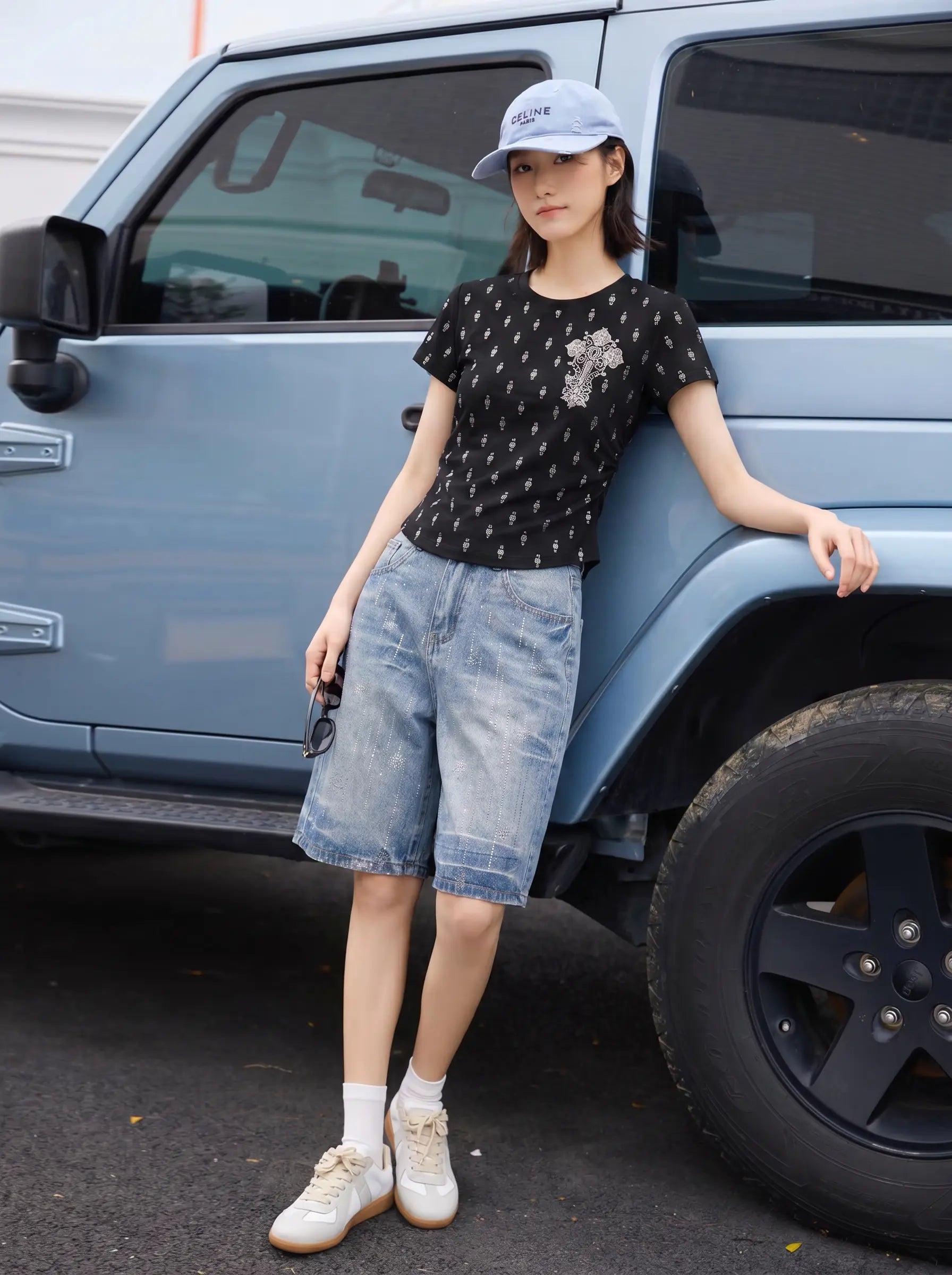 A woman stands outdoors leaning against a light blue SUV, wearing a black short-sleeve t-shirt with a small white pattern and a large white graphic on the chest, paired with light-wash denim shorts, white socks, and white sneakers. She is also wearing a light blue baseball cap. TCH Rhinestone Techno Cross Graphic Black T-Shirt