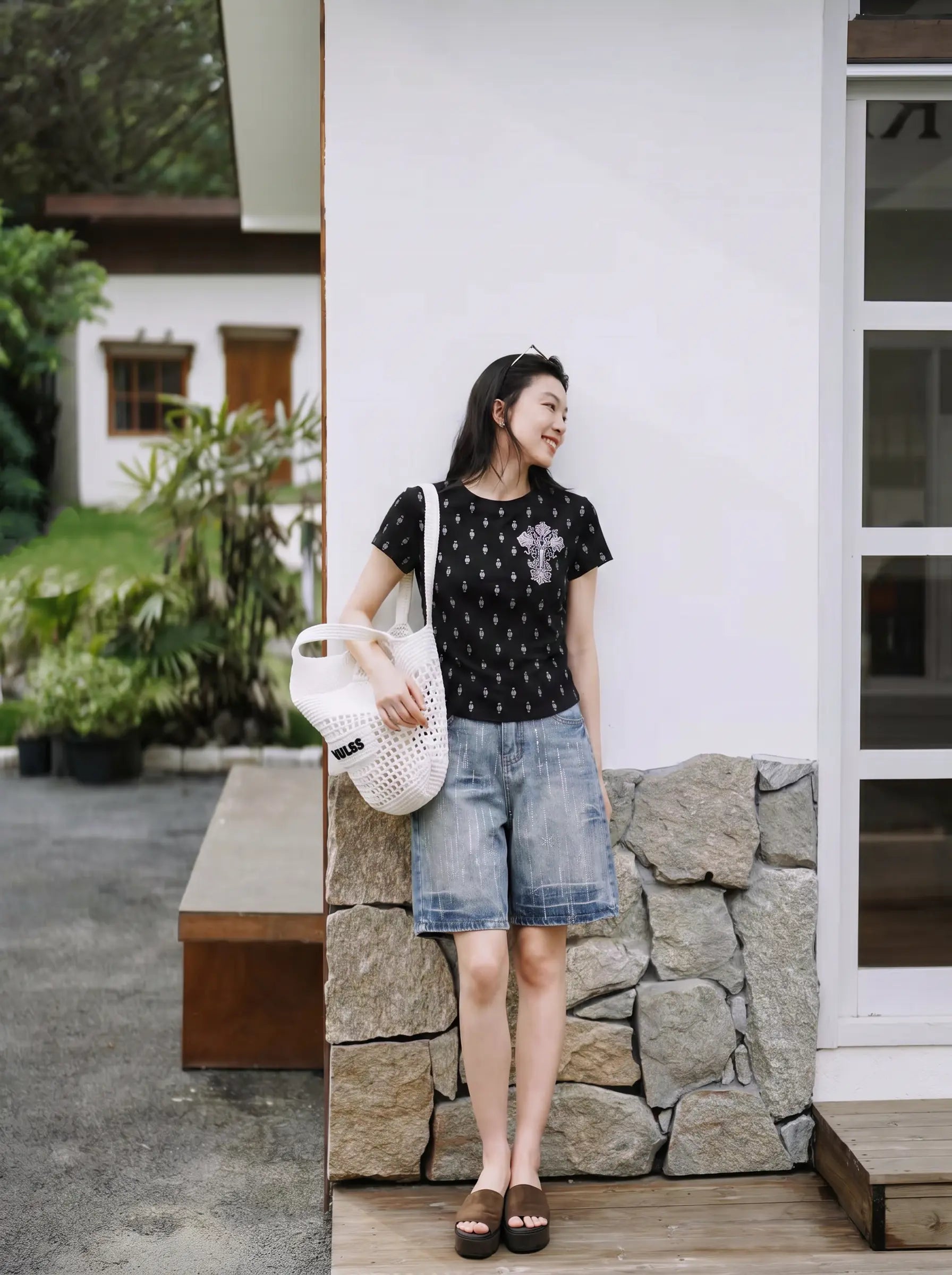 A woman stands outdoors against a white wall and stone base, wearing a black short-sleeve top with a small white floral pattern, light blue denim shorts, and dark sandals, while holding a white woven tote bag. TCH Rhinestone Techno Cross Graphic Black T-Shirt