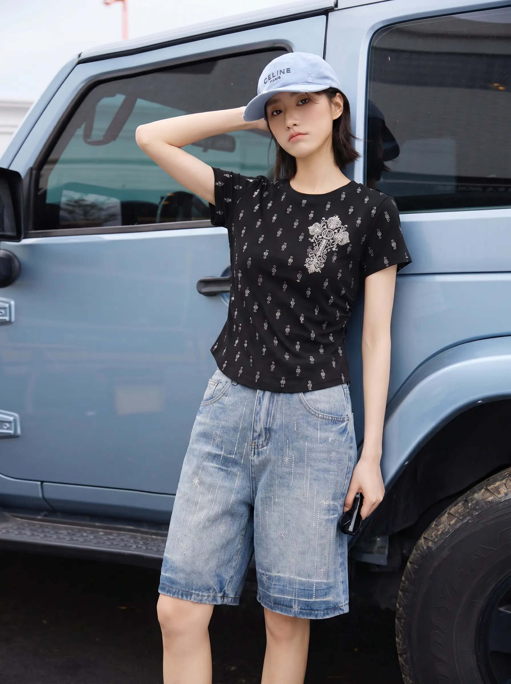 A woman stands outdoors next to a light blue SUV, wearing a fitted black short-sleeve t-shirt with a small white geometric pattern and a large silver graphic on the chest, paired with light-wash denim shorts and a light blue baseball cap. TCH Rhinestone Techno Cross Graphic Black T-Shirt
