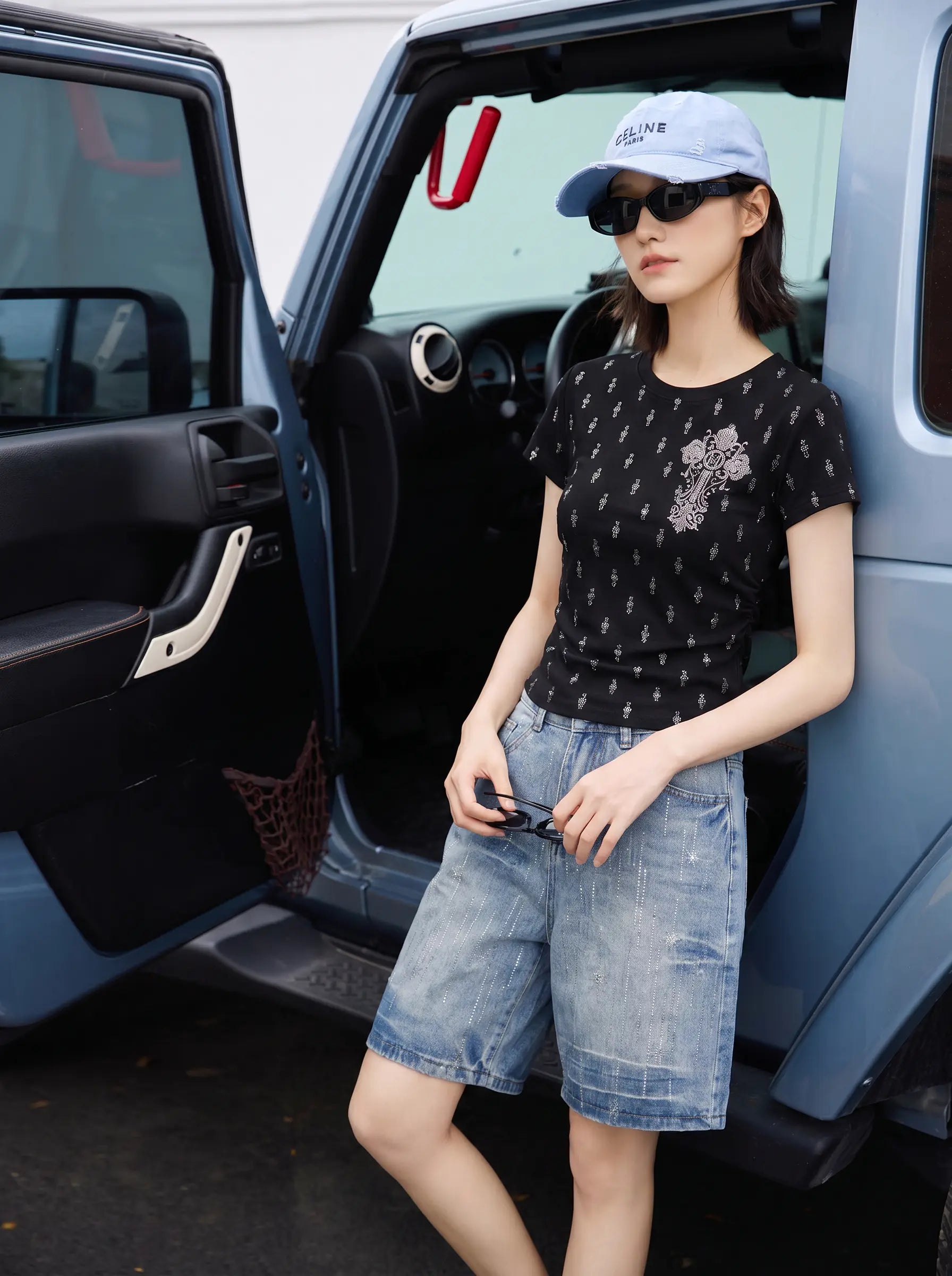 A woman stands beside an open blue vehicle door, wearing a black short-sleeve t-shirt with a small white geometric pattern and a pink graphic detail on the front, paired with light-wash denim shorts, a light blue baseball cap, and sunglasses. TCH Rhinestone Techno Cross Graphic Black T-Shirt
