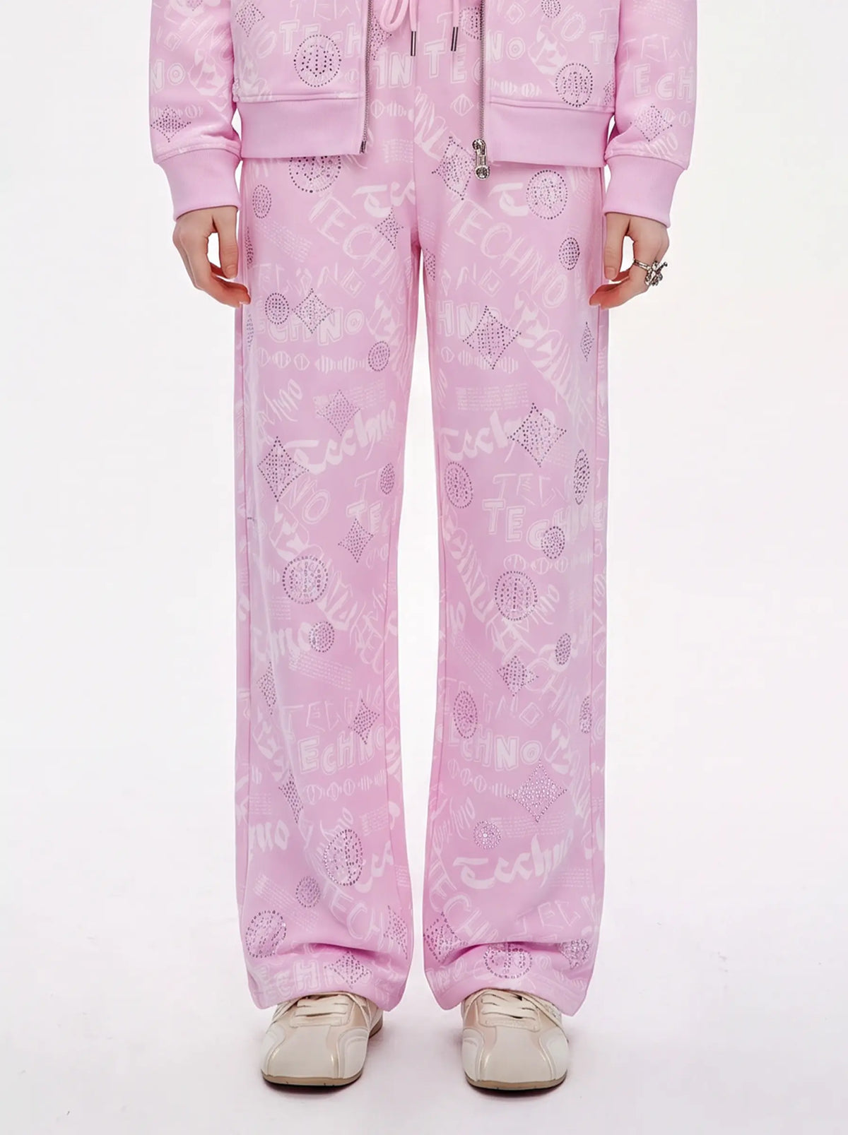 A model wearing a light pink matching tracksuit with an all-over tonal graphic print, featuring a zip-up hoodie and wide-leg sweatpants, paired with beige sneakers against a plain white background. TCH Rhinestone Techno Graphic Wide-Leg Sweatpants