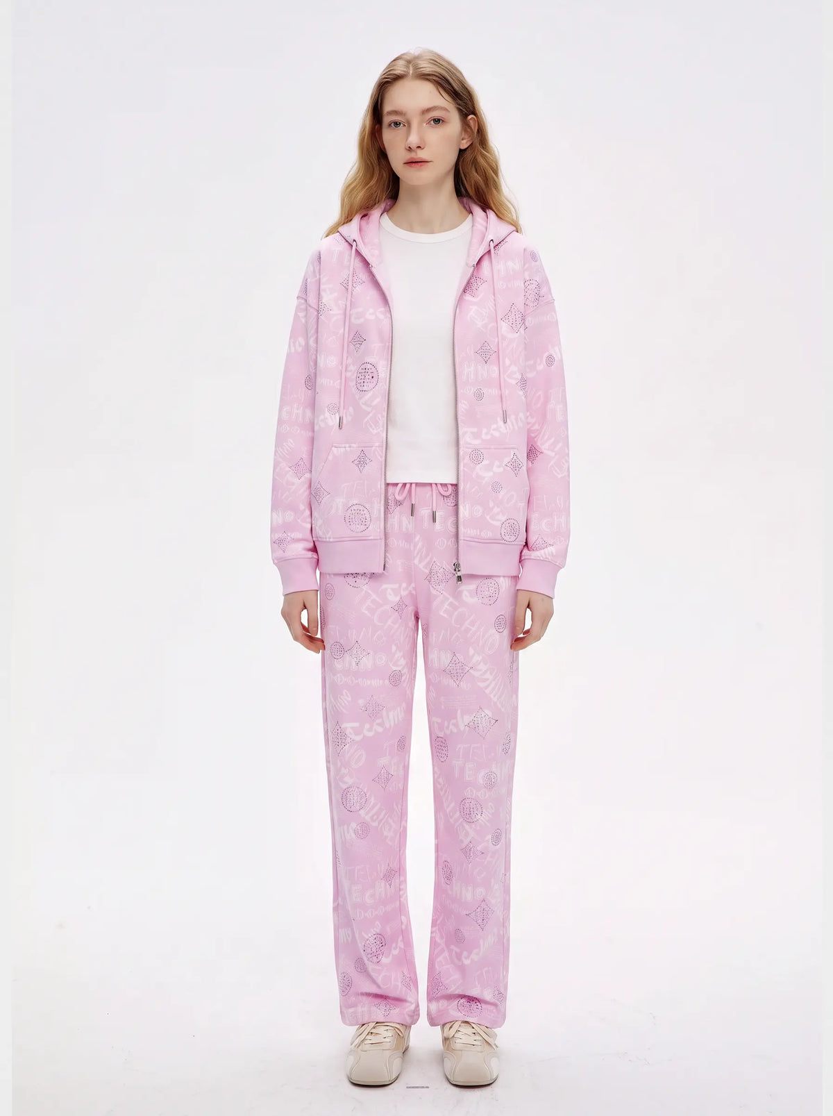 A woman stands against a plain white studio background wearing a light pink zip-up hoodie and matching jogger pants with a subtle tonal floral pattern, over a white top and light beige sneakers. TCH Rhinestone Techno Graphic Wide-Leg Sweatpants