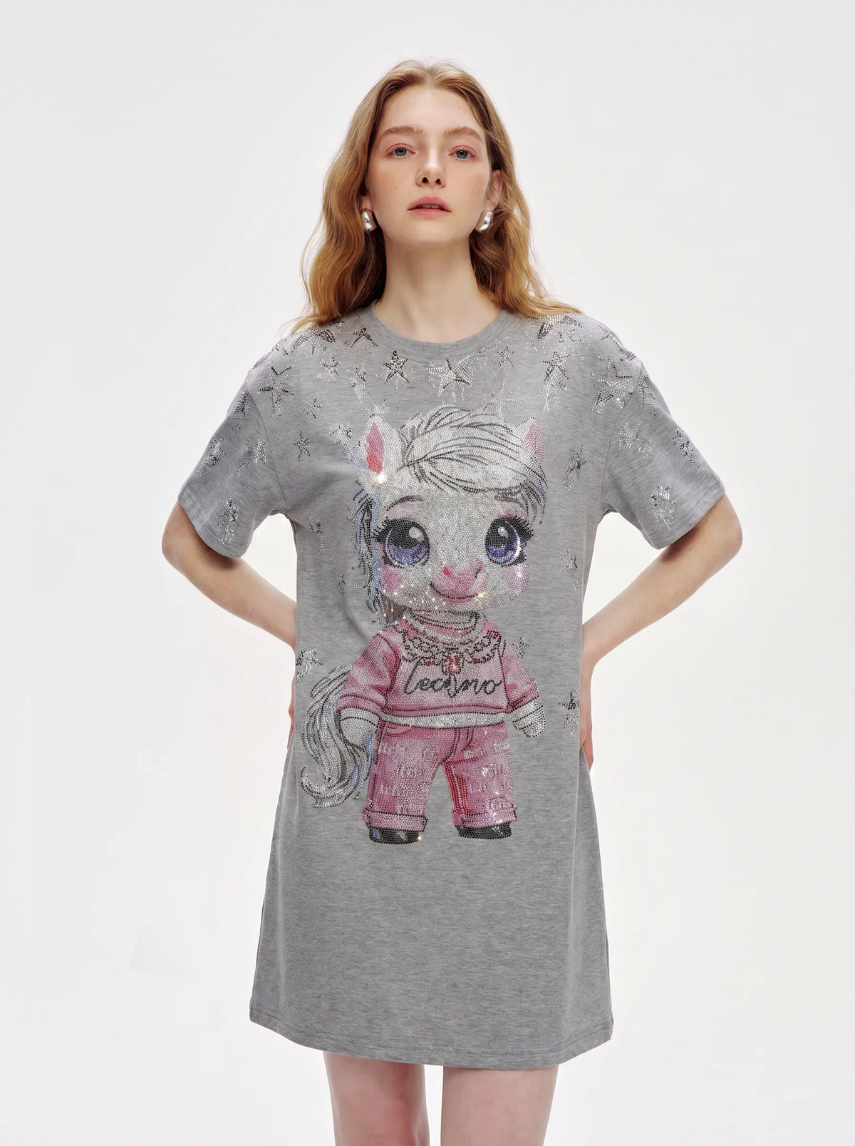 A woman modeling a heather gray short-sleeve t-shirt dress featuring a large cartoon unicorn graphic on the front in pink and white, with subtle sparkly star details across the fabric. TCH Rhinestone Techno Unicorn Graphic T-Shirt