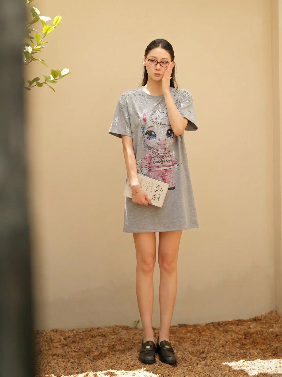 A woman wearing a gray oversized graphic t-shirt dress with a cartoon character print, holding a small book and wearing black loafers, standing against a beige wall. TCH Rhinestone Techno Unicorn Graphic T-Shirt