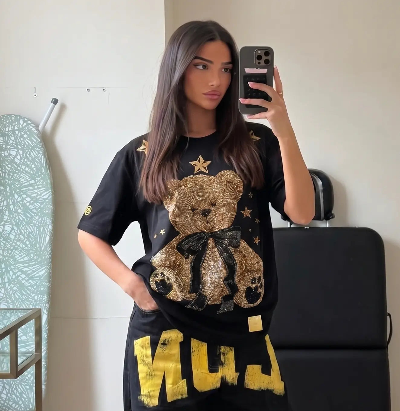 Woman wearing a black t-shirt with a teddy bear graphic and yellow text, taking a mirror selfie. TCH Rhinestone Teddy Bear Black T-Shirt