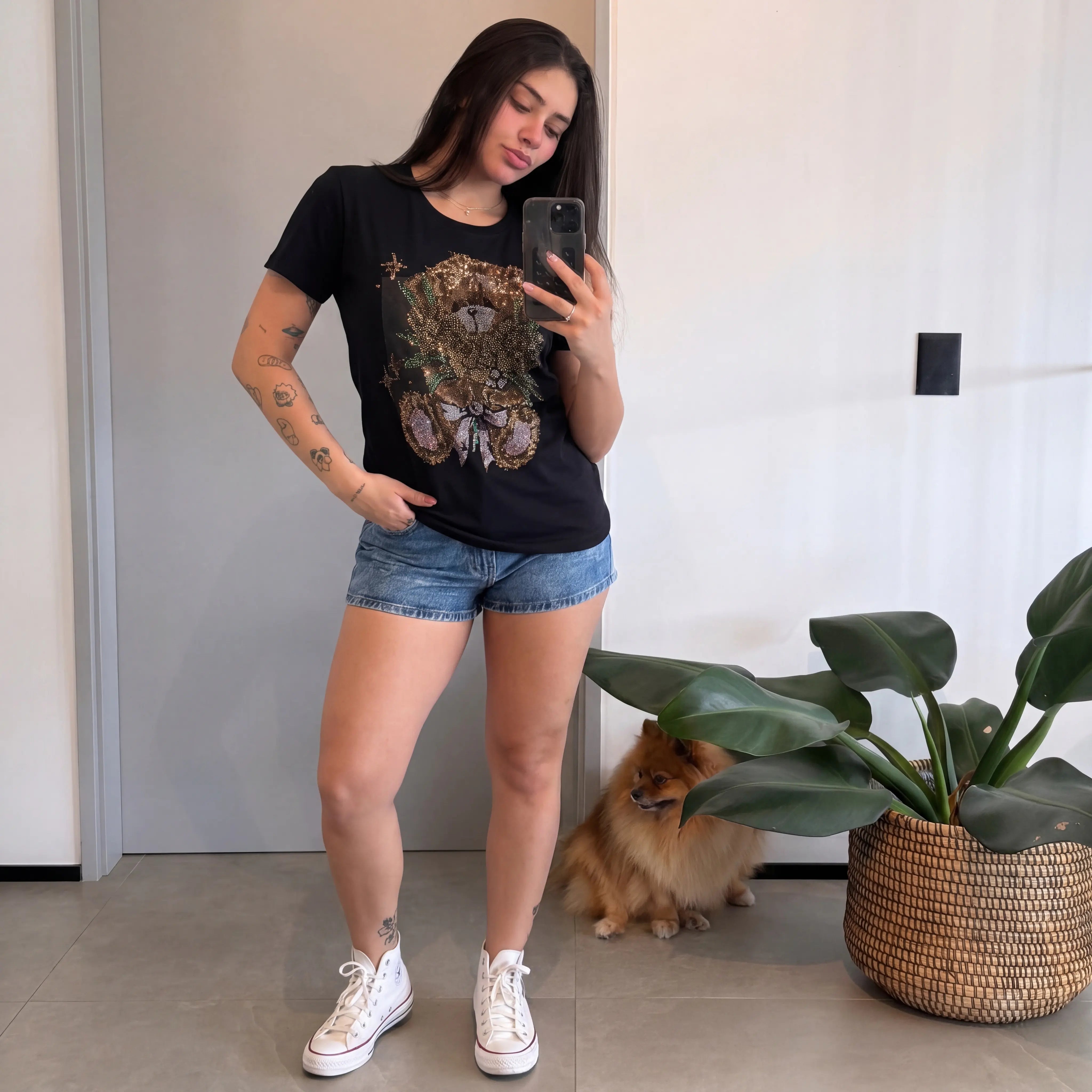 Woman taking a mirror selfie wearing a black t-shirt with a graphic design, denim shorts, and white sneakers. A dog is sitting on the floor in the background. TCH Rhinestone Teddy Bear Bouquet Black T-Shirt.