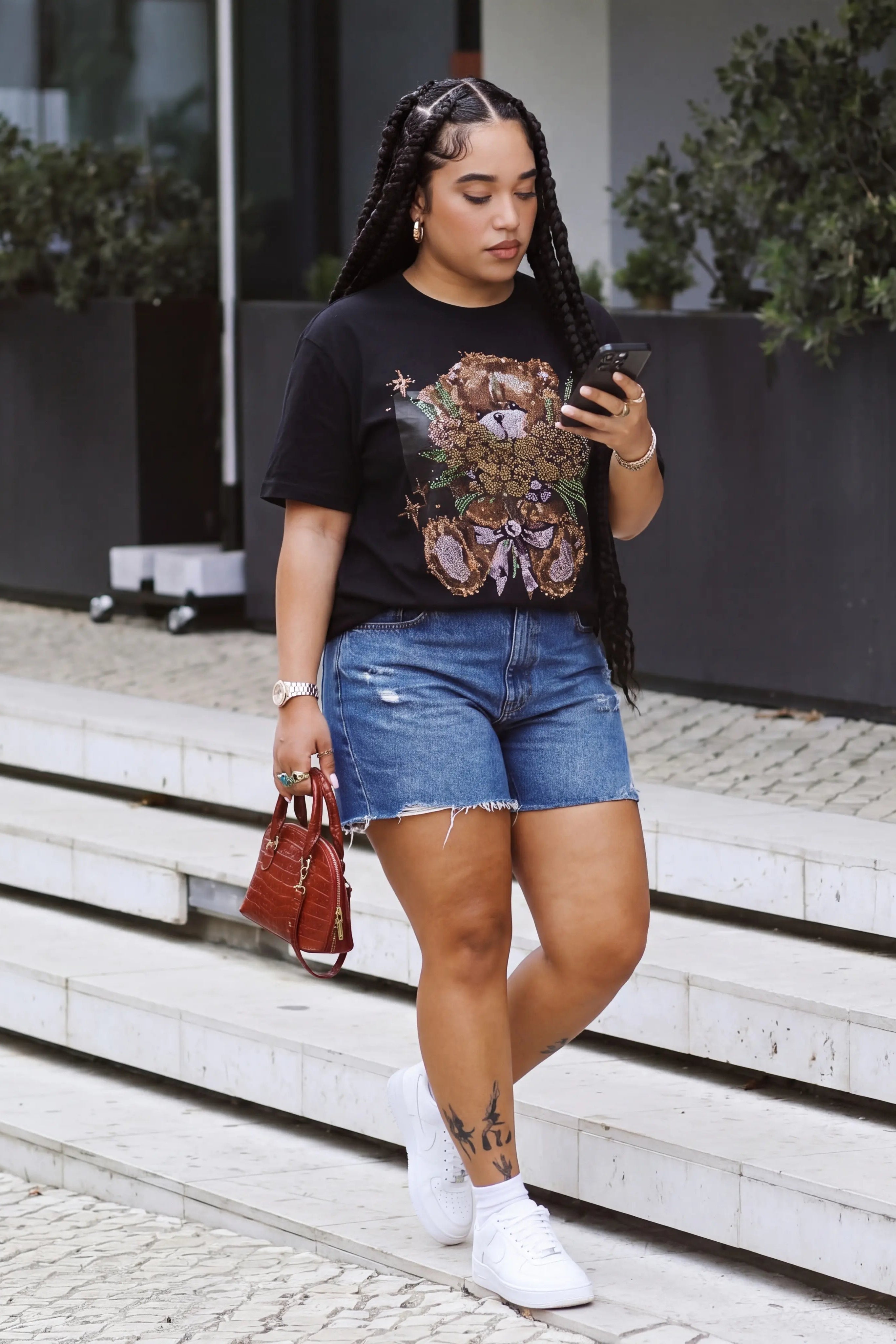 Woman standing on steps looking at her phone, wearing a black t-shirt and denim shorts. TCH Rhinestone Teddy Bear Bouquet Black T-Shirt.