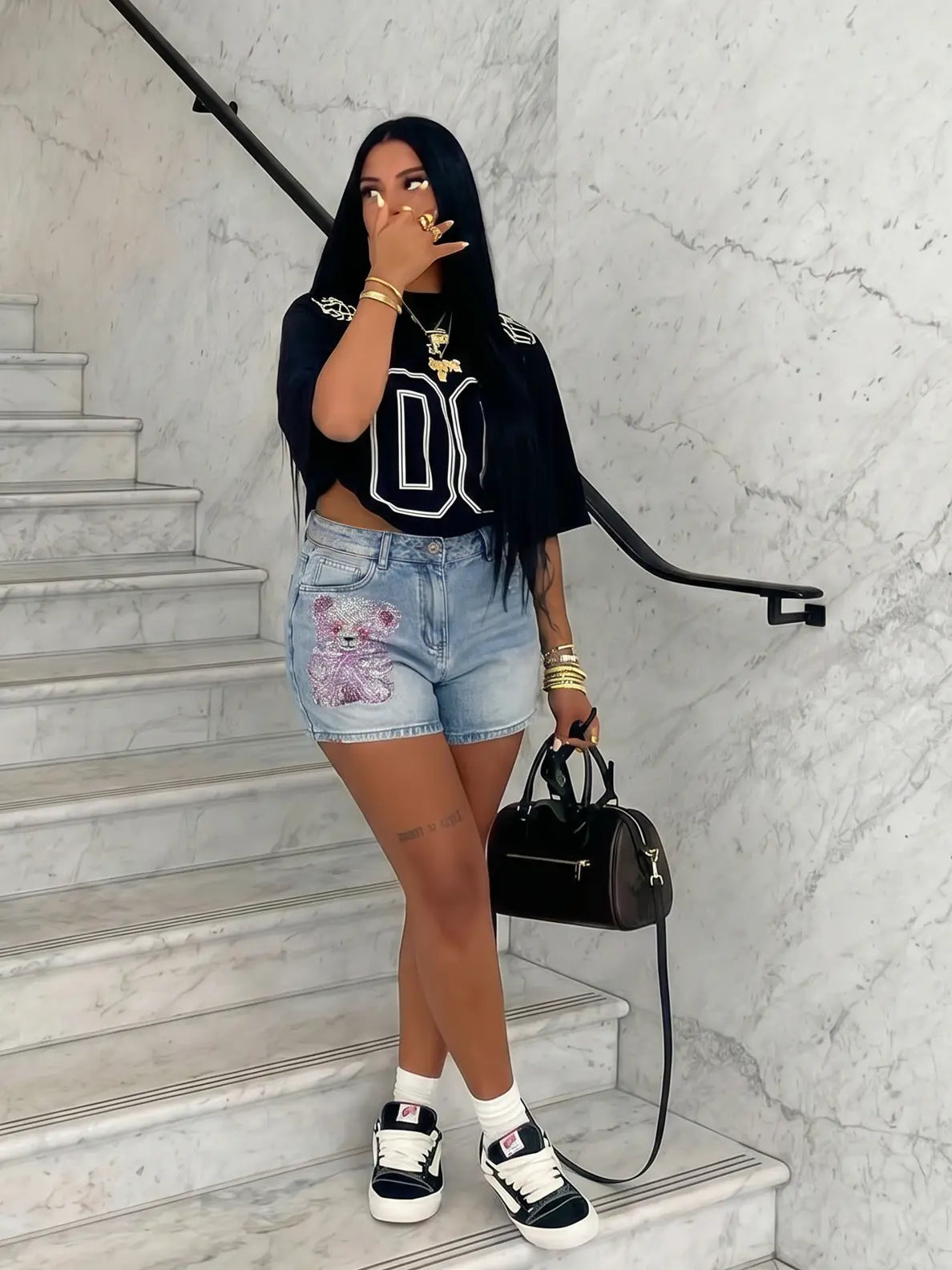 A woman posing on a marble staircase wearing a black cropped t-shirt with a white graphic, light-wash denim shorts with a pink graphic, black and white sneakers, and holding a black handbag. TCH Rhinestone Teddy Bear Bow Denim Shorts