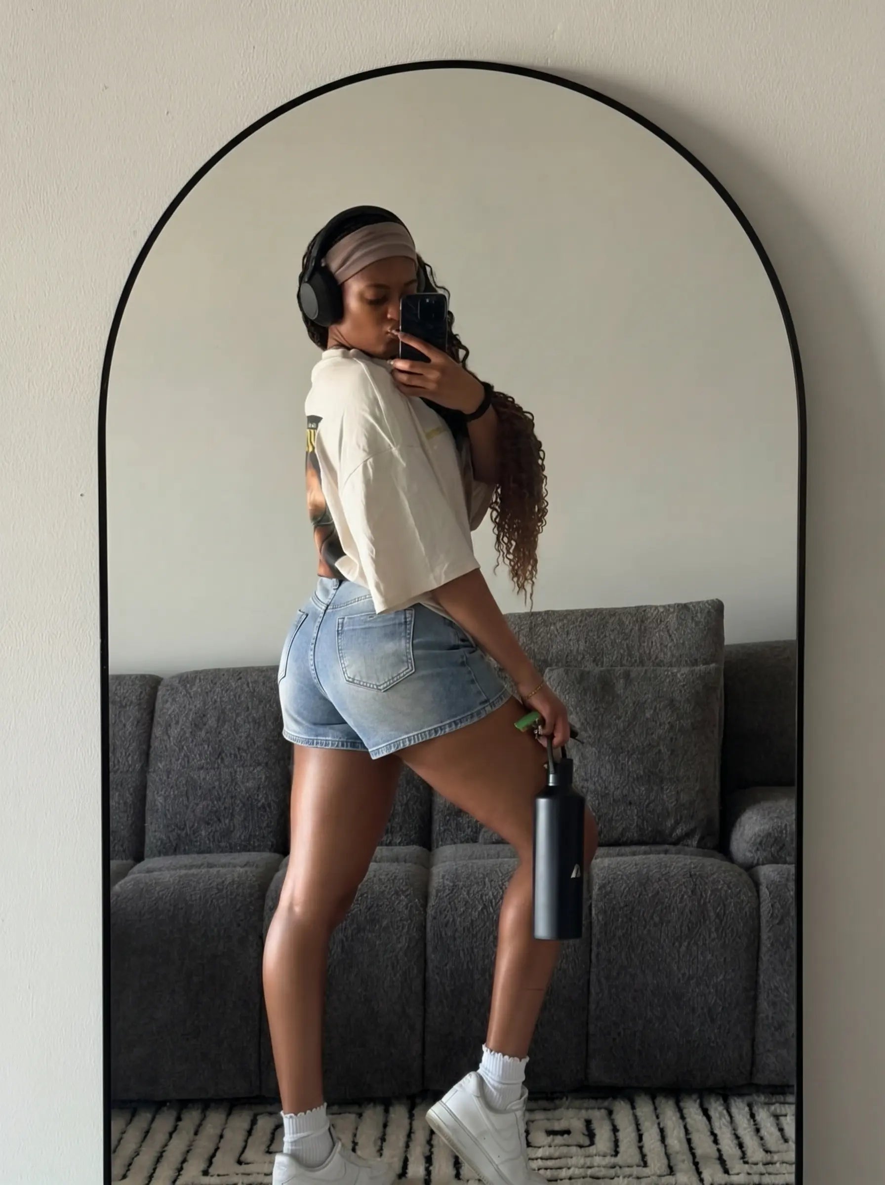 A woman takes a mirror selfie in a living room, wearing a white cropped graphic t-shirt, light-wash denim shorts, white socks, and white sneakers, while holding a black water bottle. She is positioned in front of a gray sofa and a large arched floor mirror. TCH Rhinestone Teddy Bear Bow Denim Shorts
