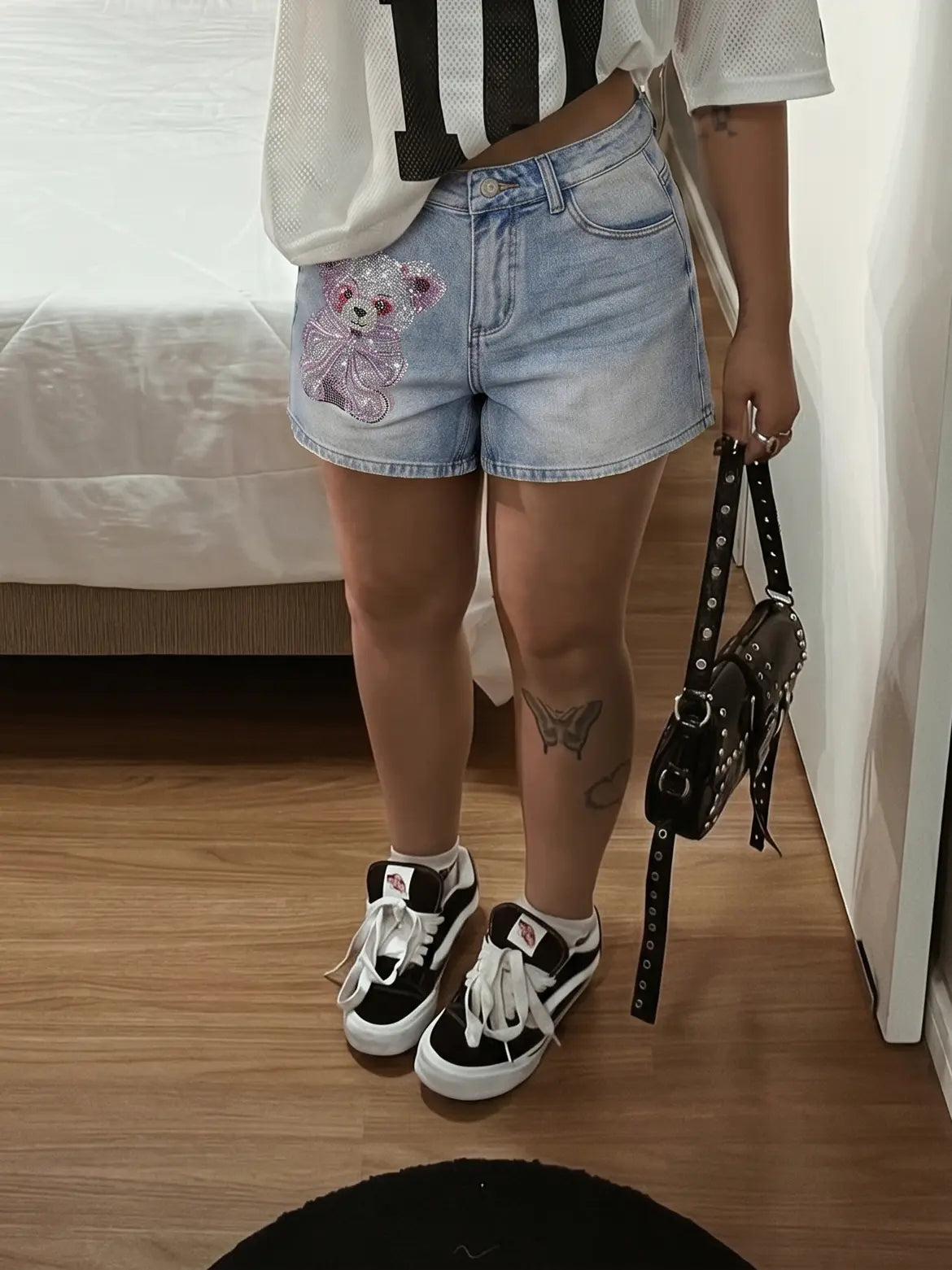 A person standing indoors wearing light-wash denim shorts with a pink teddy bear patch, a white cropped top with black stripes, black-and-white sneakers, and holding a black studded shoulder bag. TCH Rhinestone Teddy Bear Bow Denim Shorts