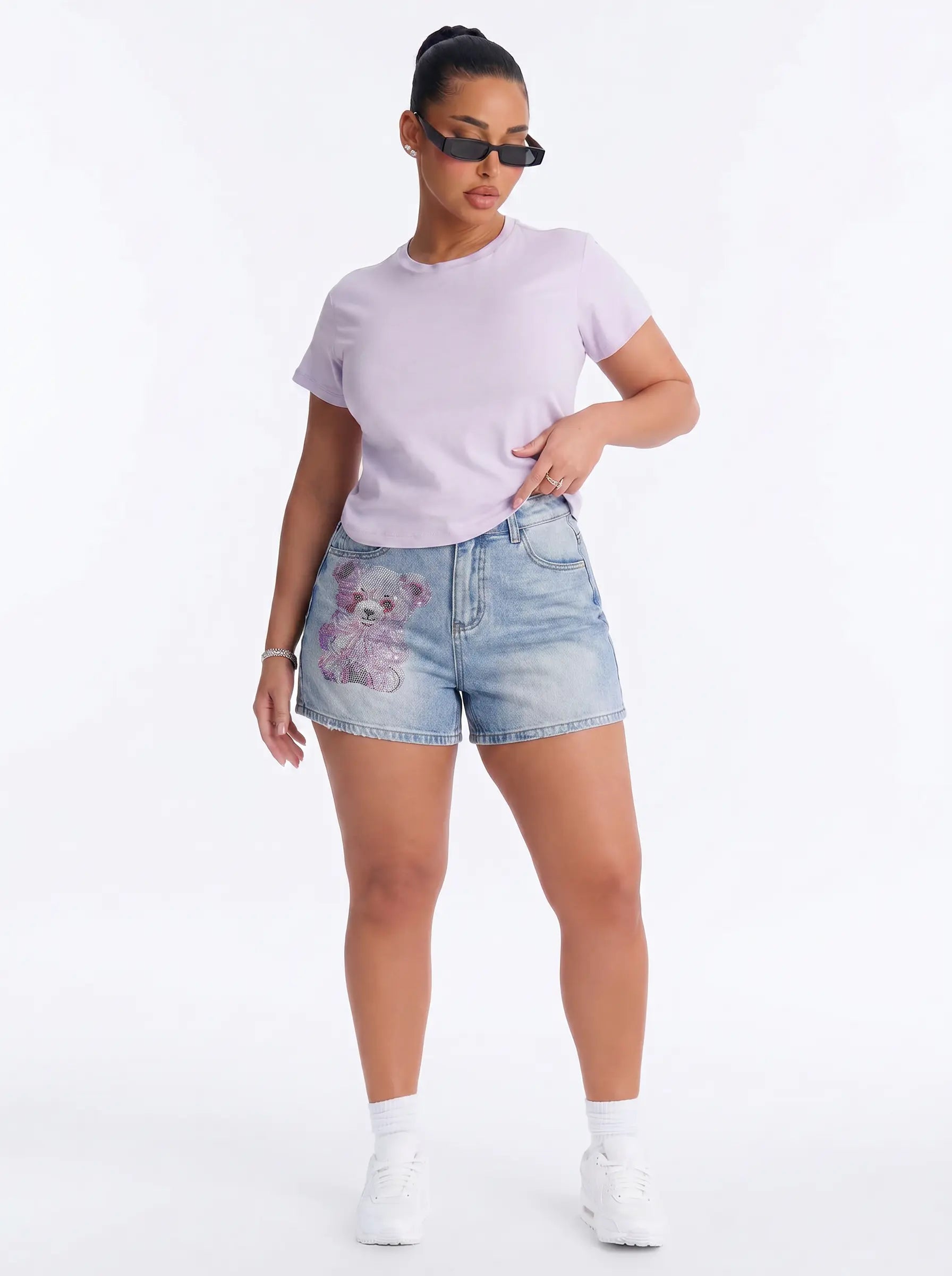 A woman modeling a light purple short-sleeve t-shirt and light-wash denim shorts with a purple teddy bear patch on the left leg, styled with white sneakers and black sunglasses against a white studio background. TCH Rhinestone Teddy Bear Bow Denim Shorts