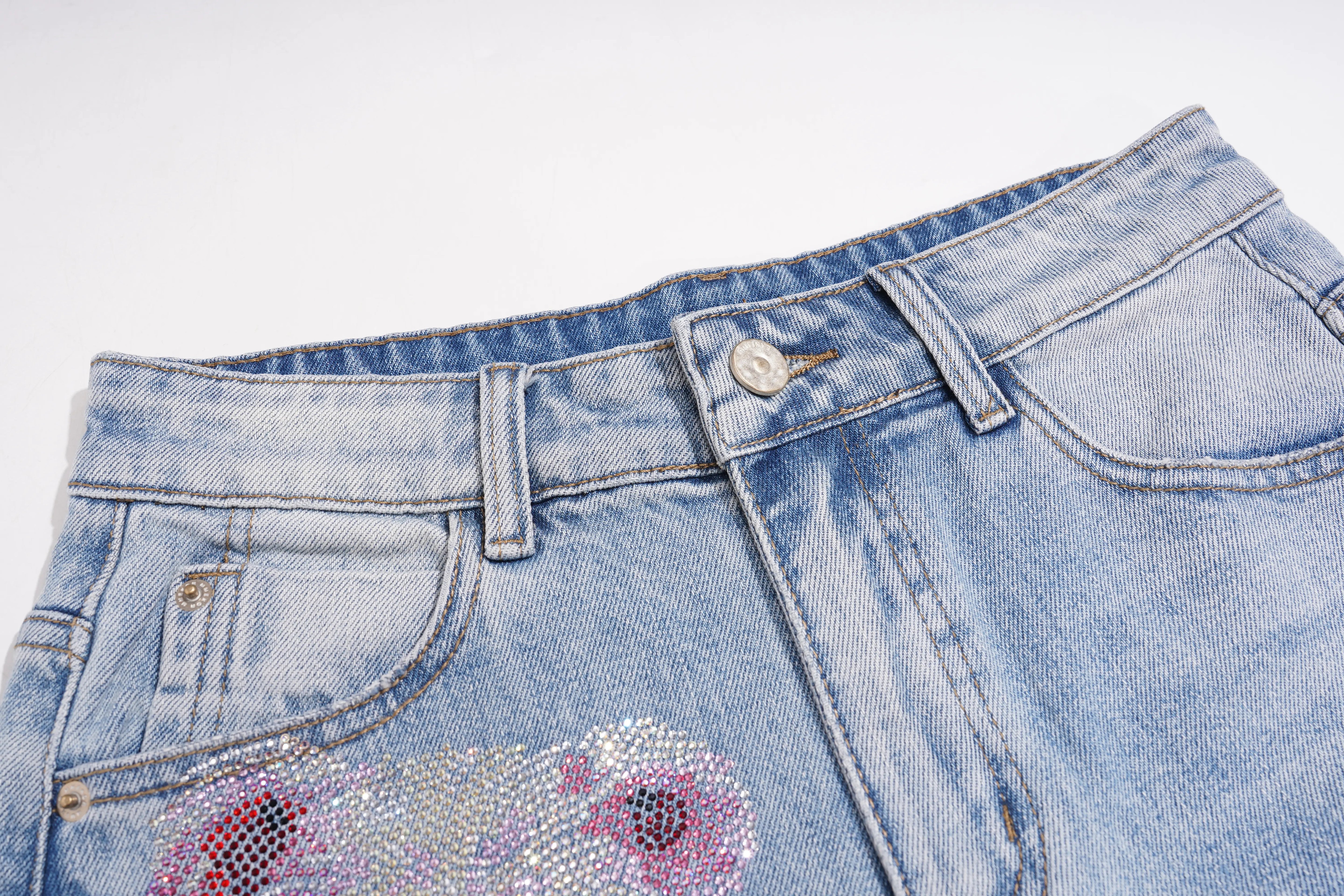 Close-up of light blue denim jeans showing the waistband, belt loops, front pocket, and a decorative sequined floral embellishment on the front right leg. TCH Rhinestone Teddy Bear Bow Denim Shorts