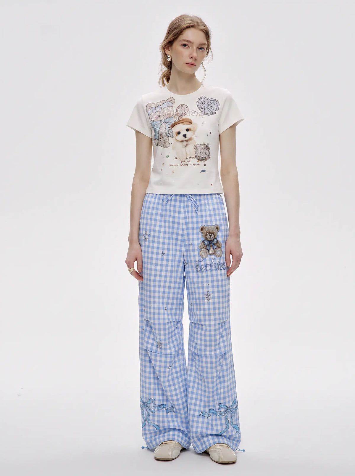 A model stands against a light gray studio background wearing a cream graphic T-shirt with cartoon animals and blue-and-white gingham wide-leg pants with a teddy bear patch on the pocket. TCH Rhinestone Teddy Bear Bow Gingham Wide-Leg Pants