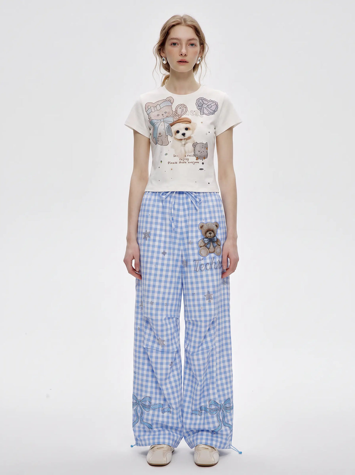A model stands against a light gray studio background wearing a cream graphic T-shirt with teddy bear illustrations and blue-and-white gingham wide-leg pants with a teddy bear patch. TCH Rhinestone Teddy Bear Bow Gingham Wide-Leg Pants