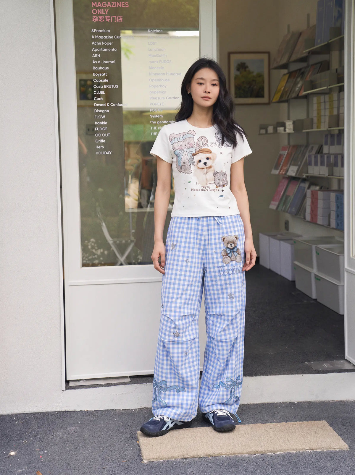 A woman stands in front of a shop doorway wearing a white graphic T-shirt with a cartoon dog and blue-and-white gingham wide-leg pants with a teddy bear patch. TCH Rhinestone Teddy Bear Bow Gingham Wide-Leg Pants