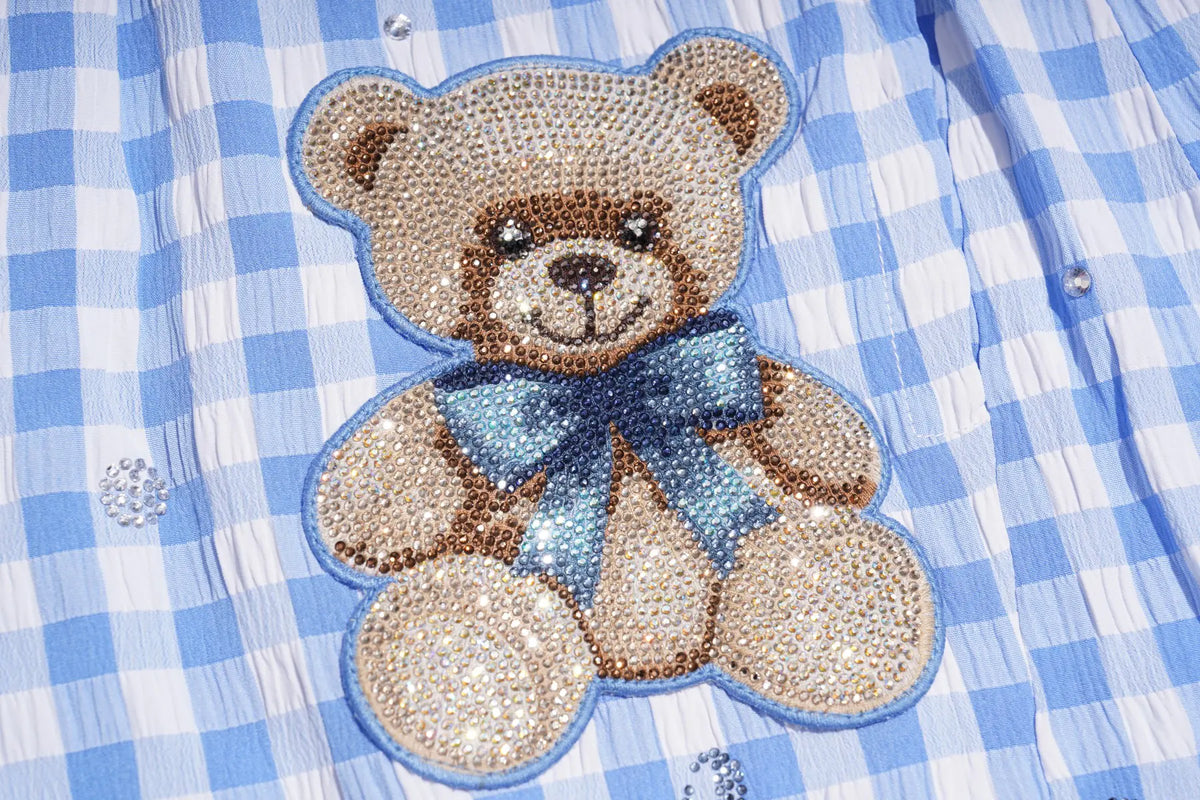 A close-up of a blue and white gingham shirt featuring a large, beaded teddy bear applique on the chest. The teddy bear is beige with brown details and a sparkling blue bow, outlined in blue beads. TCH Rhinestone Teddy Bear Bow Gingham Wide-Leg Pants
